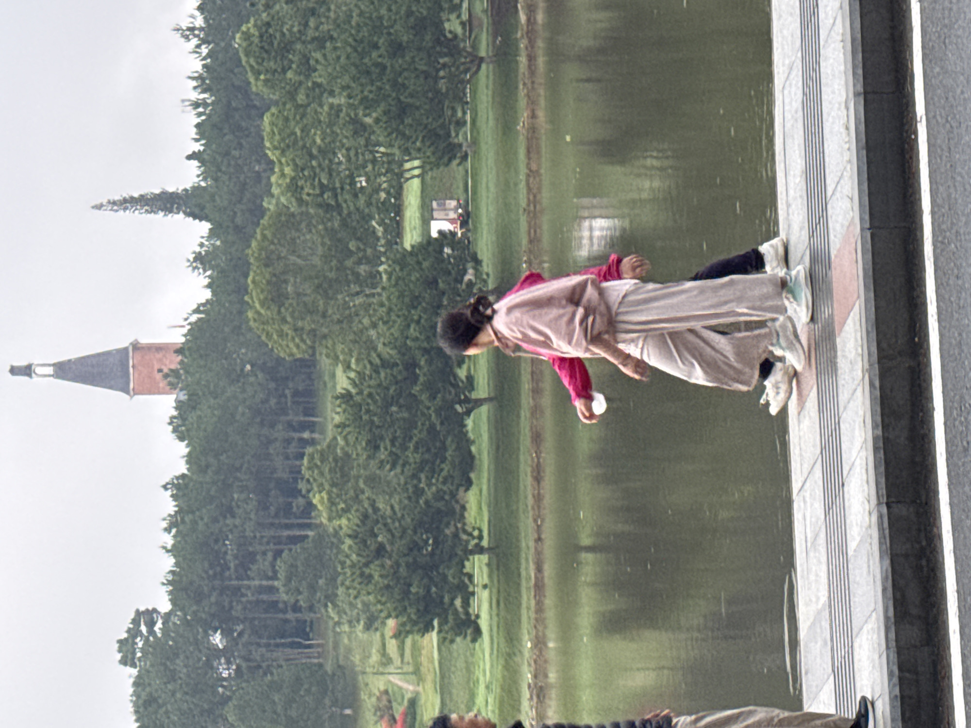 The image depicts Xuan Huong Lake in Da Lat, Vietnam. In the foreground, two individuals are walking along a paved pathway bordering the lake. The person on the left is partially visible, wearing light-colored pants and a dark jacket. The person on the right is more prominently featured, wearing a light pink top and loose-fitting beige pants, with a white cup in their hand. They are walking away from the viewer. The lake itself occupies the middle ground, its surface reflecting the overcast sky and the lush green trees that line its banks. In the background, a dense forest of pine trees is visible, with a distinctive red brick tower topped with a dark spire rising above the trees. The overall atmosphere is subdued, likely due to the cloudy weather.