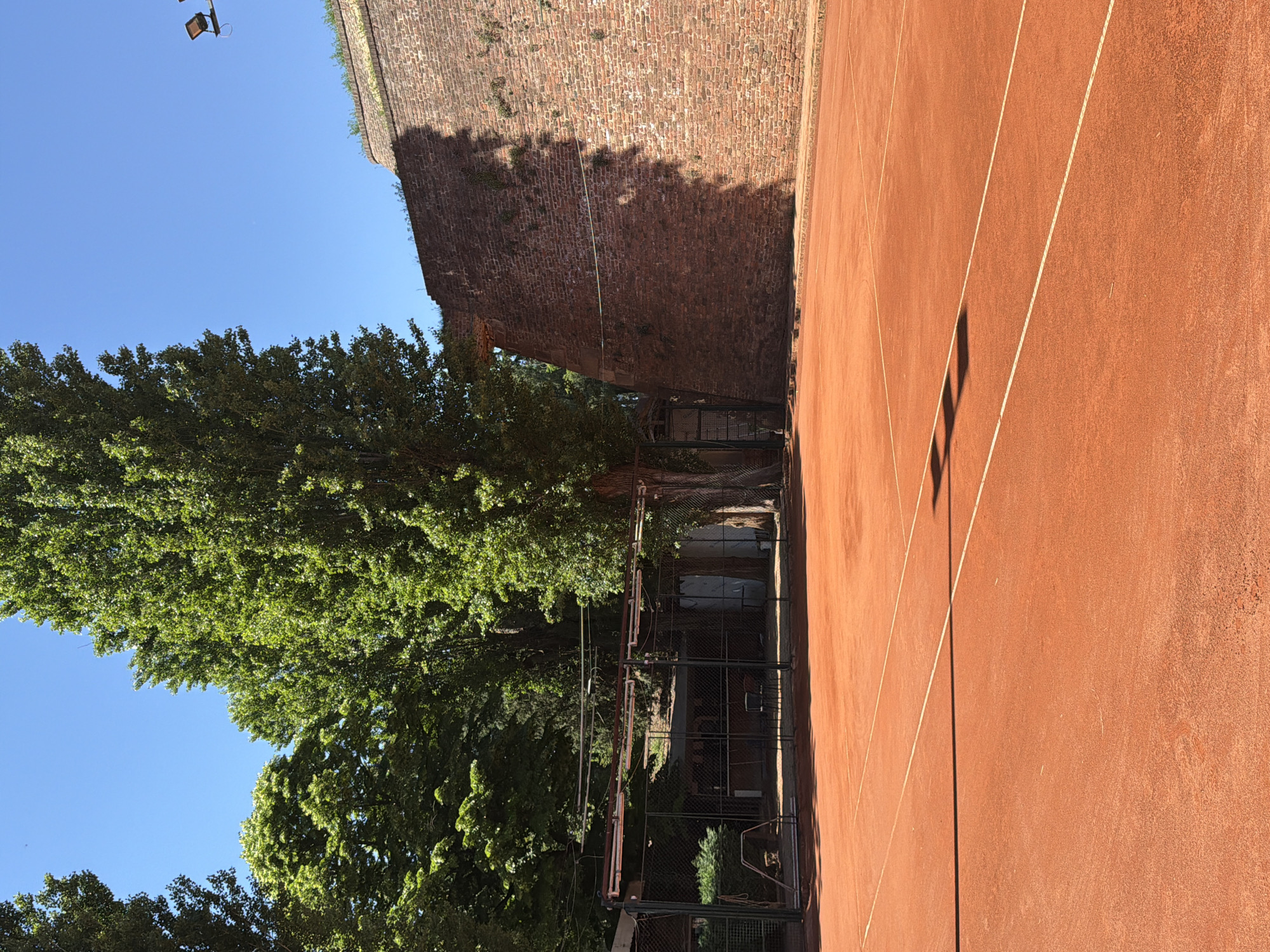 The image depicts a tennis court located in the Kalemegdan Fortress in Belgrade, Serbia. The court has a red clay surface with white lines marking the boundaries. Adjacent to the court is a high brick wall of the fortress, with vegetation growing on its surface. A tree with green foliage is positioned beside the wall. A metal fence is also visible between the court and the tree. The sky is blue.