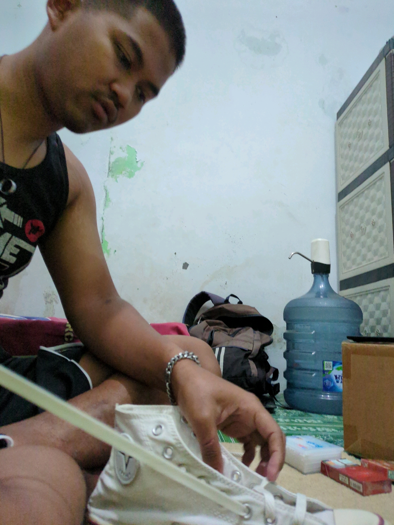 A young man is sitting on a mat in a room. He is wearing a black tank top and shorts. He is holding a white sneaker and appears to be lacing it up. To his left, there is a backpack and a large blue water jug with a pump on top. In the background, there is a white wall with some peeling paint and a brown cardboard box. There is also a pack of cigarettes on the mat. The scene is indoors, and the lighting appears to be dim.