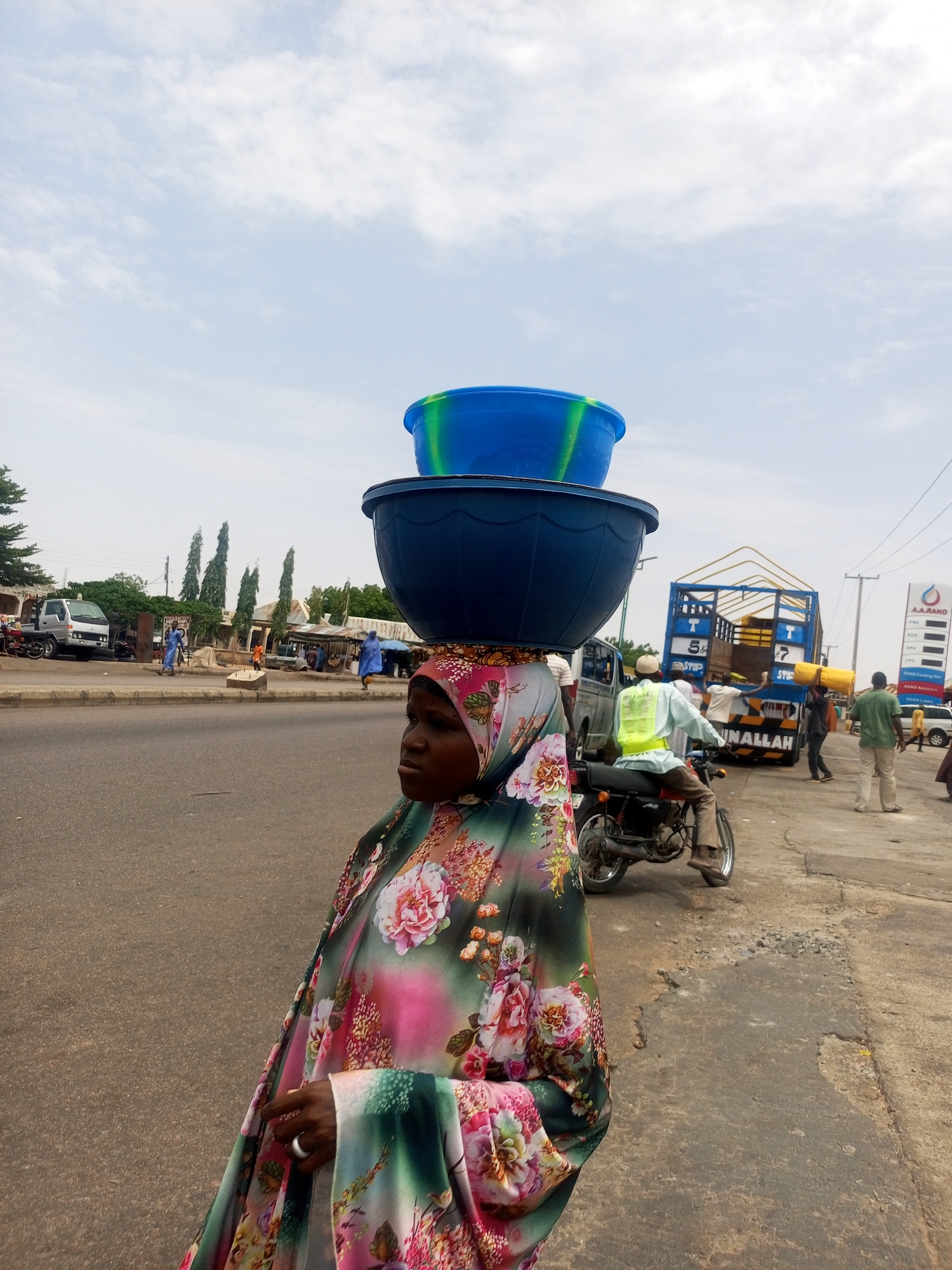 The image depicts a woman in Gombe, Nigeria, carrying two blue plastic tubs stacked on her head. She is wearing a floral print hijab and dress. The street scene includes a motorcycle with a rider wearing a reflective vest, a truck being loaded with yellow bags, a gas station sign in the background, and other pedestrians. The road is paved but shows some signs of wear.