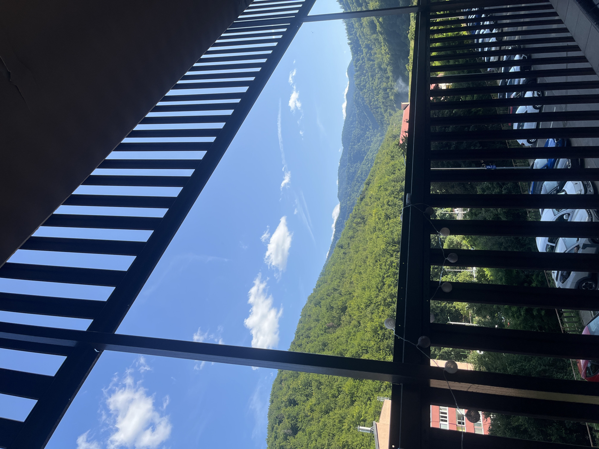 The image depicts a view from a balcony overlooking a lush, green mountainside under a clear blue sky with scattered white clouds. The foreground features a dark railing with string lights. In the midground, a red-brick building with white windows is visible on the left, followed by a parking lot filled with various cars. The background is dominated by rolling, tree-covered mountains, suggesting a location in a mountainous region. Given the context of Brasov, Romania, this scene likely captures a view from an apartment or hotel in a neighborhood situated near the foothills of the Carpathian Mountains. The overall atmosphere is serene and natural, with a hint of urban presence from the buildings and cars.