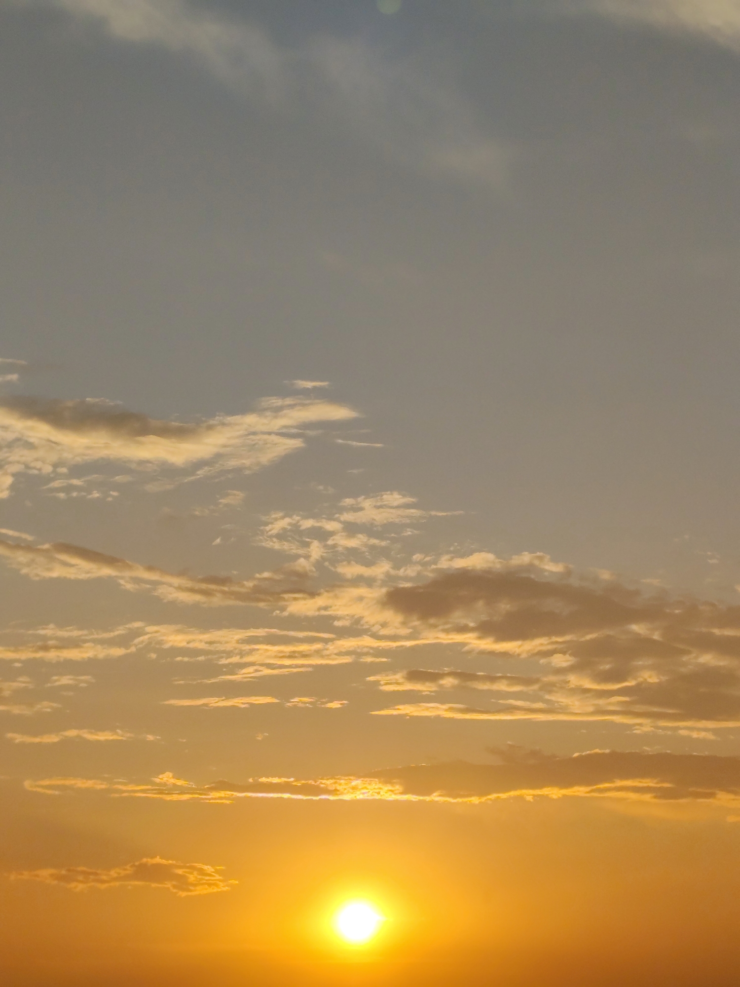 The image captures a vibrant sunset over Приишим ауылдық округі, Kazakhstan. The sky is a gradient of warm colors, transitioning from a pale blue at the top to a deep orange and yellow near the horizon. Wispy clouds are scattered across the sky, some tinged with gold by the setting sun. The sun itself appears as a bright, luminous orb on the right side of the frame, casting a warm glow over the scene. The lower portion of the image is dominated by the intense light of the sunset. There are no people or discernible structures visible in this landscape photograph.
