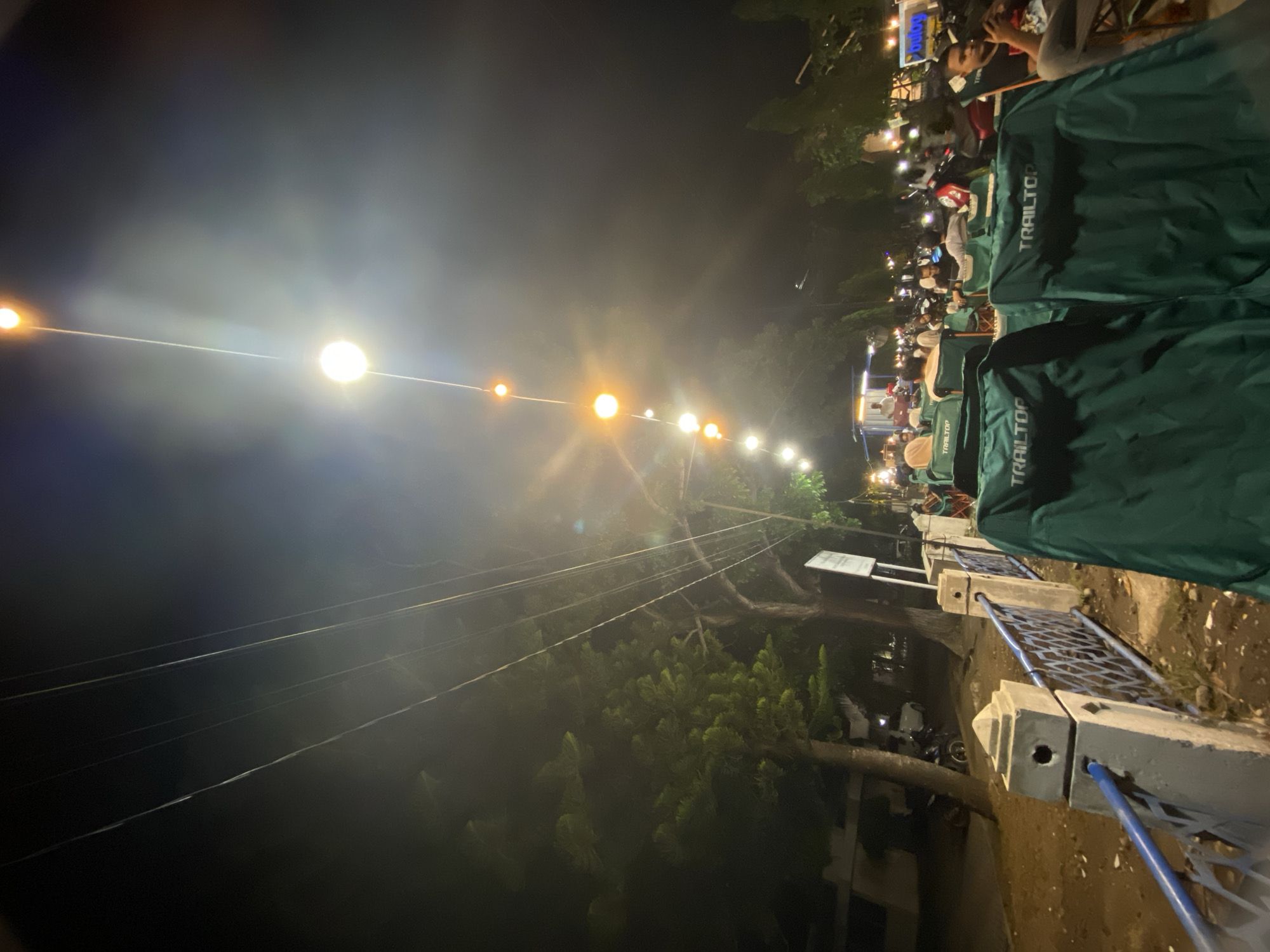 This is a nighttime, medium-shot photograph taken at a bustling outdoor gathering, likely a food court or event space, in Lhokseumawe, Indonesia. The scene is illuminated by strings of lights, with a prominent line of warm-toned bulbs stretching diagonally across the upper portion of the frame, casting a soft glow. Several people are visible, seated in green camping chairs labeled "TRAILTOPTOP" in the foreground. These chairs face away from the camera, suggesting an audience observing something out of frame. Behind the chairs, more people are scattered, some on motorcycles, indicating it's a public space. The background includes trees, a blue and white fence, and the dark night sky. The overall atmosphere is social and lively, with the lights creating a festive ambiance.
