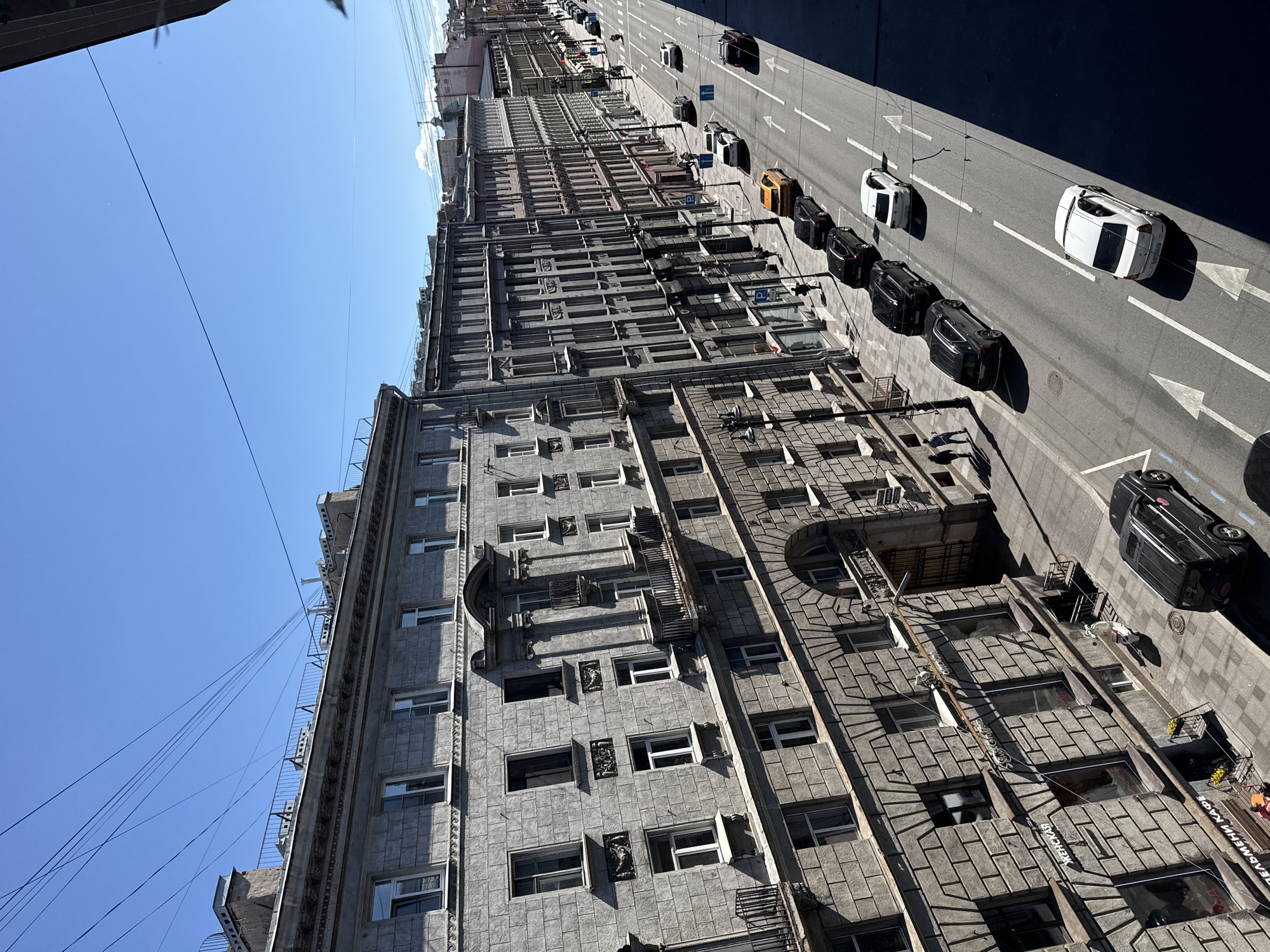 The image captures a street scene in a city, likely St. Petersburg, Russia, given the architectural style. The perspective is from a high vantage point, looking down a wide street lined with multi-story buildings. The buildings are constructed of stone and feature a classical architectural design with numerous windows, balconies, and decorative elements. Some buildings have storefronts at the ground level.
The street itself is paved and marked with traffic lanes and directional arrows. Several cars are visible, parked along the side of the road and driving in the lanes. These include a black SUV, a white sedan, and an orange sedan, among others. Pedestrians are seen walking on the sidewalk. The sky is clear and blue, indicating a sunny day. Overhead, numerous power lines crisscross the sky. The overall scene depicts a bustling urban environment on a bright, clear day.