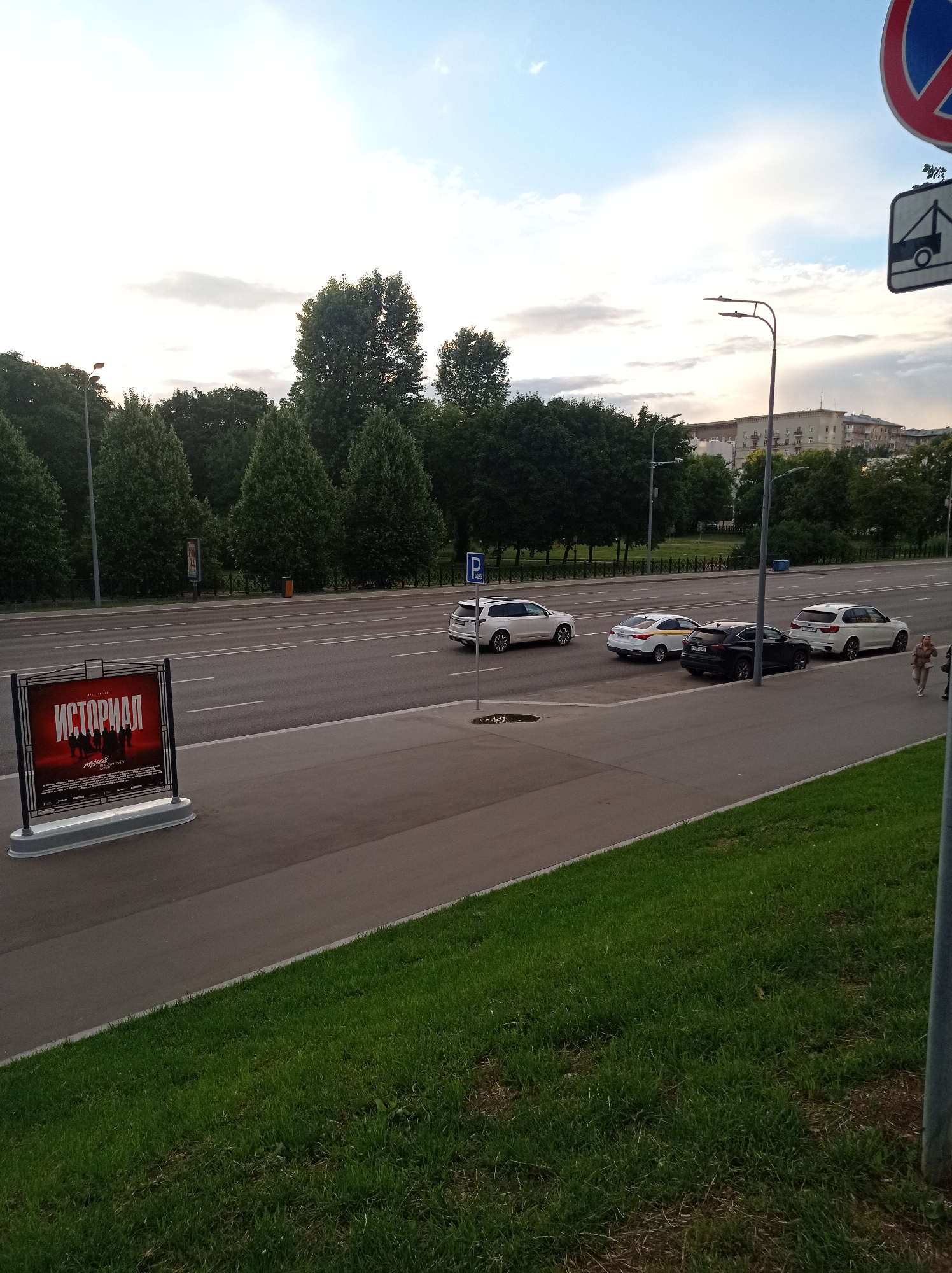 The image shows a street scene in Moscow, Russia, with a road and sidewalk in the foreground. On the sidewalk, there is a large red billboard with Russian text that reads "ИСТОРИМАЛ". Several cars are driving or parked on the road, including a white SUV, a black sedan, and a white station wagon. A woman is walking on the sidewalk in the background. The street is lined with trees and buildings, and the sky is partly cloudy. There are also streetlights and traffic signs visible. The billboard appears to be promoting a movie or event. The scene is captured during daylight hours.