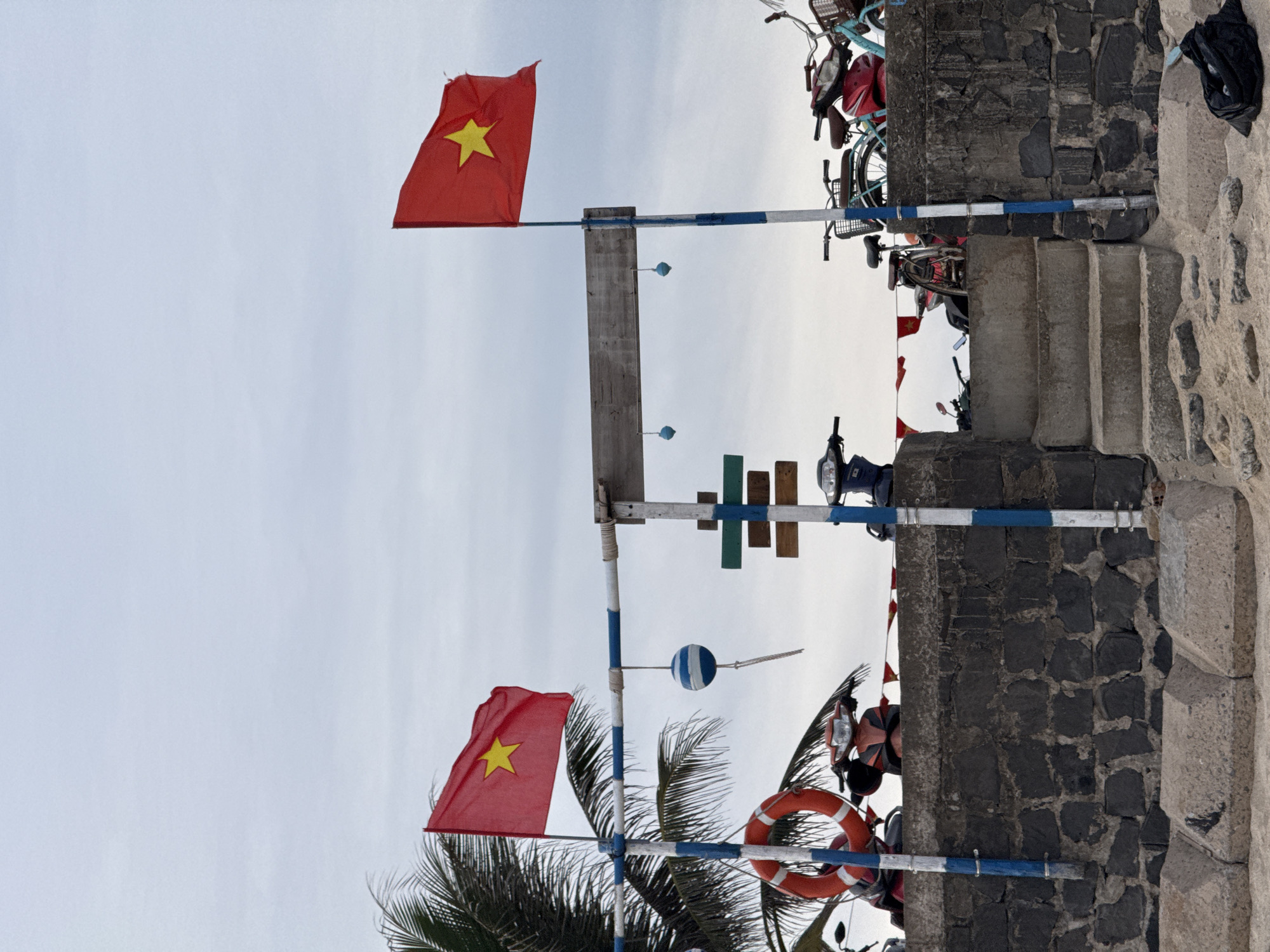 This image depicts a scene in Lý Sơn Special Administrative Region, Vietnam, featuring two Vietnamese flags flying from poles. The flags are red with a yellow star in the center. In the foreground, a rough stone wall forms a barrier, with concrete steps leading up to a sandy area. Several motorbikes and bicycles are parked behind the wall. A life preserver ring is visible hanging from a pole on the left. The sky is overcast. The overall environment suggests a coastal or beachfront location, likely a public area given the presence of flags and parked vehicles. There are no people visible in the frame.