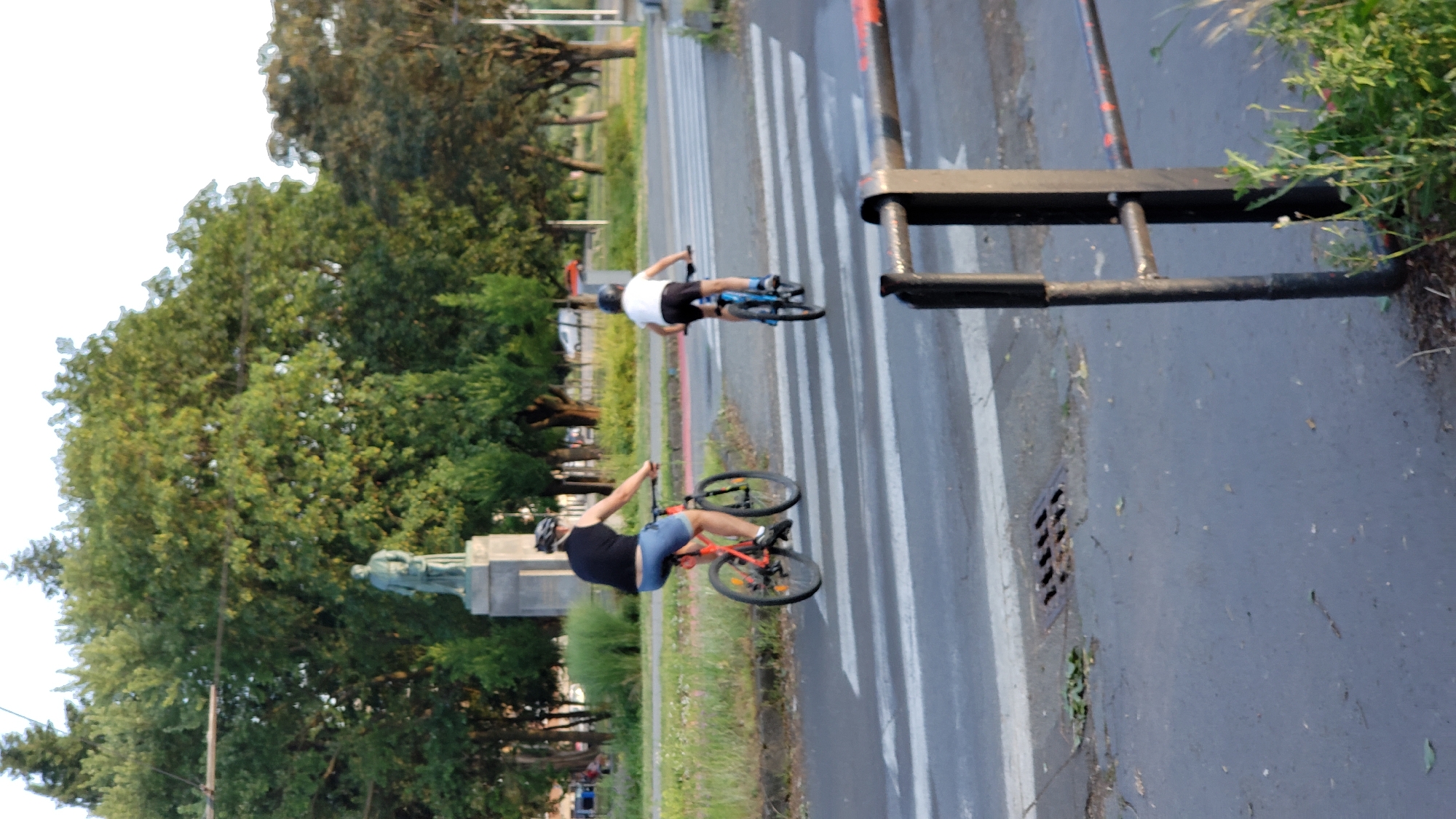 Two people are riding bicycles across a crosswalk. The person in the foreground is wearing a black tank top, blue shorts, and a helmet, and is riding a red bicycle. They are leaning back on their bicycle, with their front wheel slightly lifted. The person in the background is wearing a white t-shirt, black shorts, and a helmet, and is riding a blue bicycle. They appear to be cycling normally. The scene is set on a paved road with white crosswalk markings. Trees are visible on the left side of the road, and a black metal barrier is on the right. The lighting suggests it is daytime, likely late afternoon or early evening due to the angle of the sun. There are no visible text elements in the image. The overall mood seems casual and recreational. The presence of a crosswalk and a barrier suggests an urban or park setting.