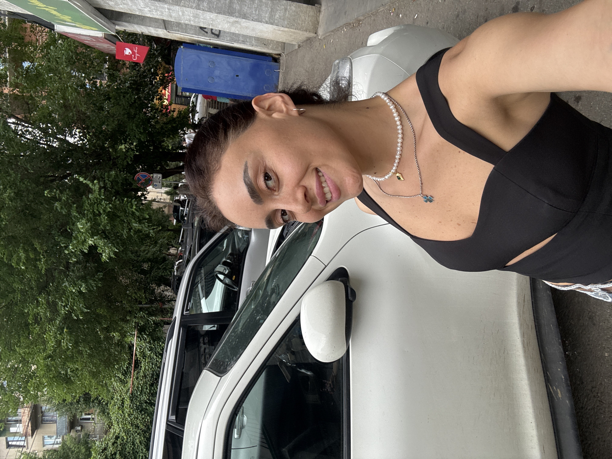 The media file shows a woman in a black top and pearl necklace standing next to a white car. She is taking a selfie, with the camera angled downwards. The woman has dark hair, is smiling, and appears to be in good spirits. In the background, there are trees, buildings, and a blue trash receptacle. A red flag with a white logo is visible on a pole. The scene appears to be outdoors, possibly on a street in Tbilisi, Georgia. The time of day is unclear, but the lighting suggests it is daytime.