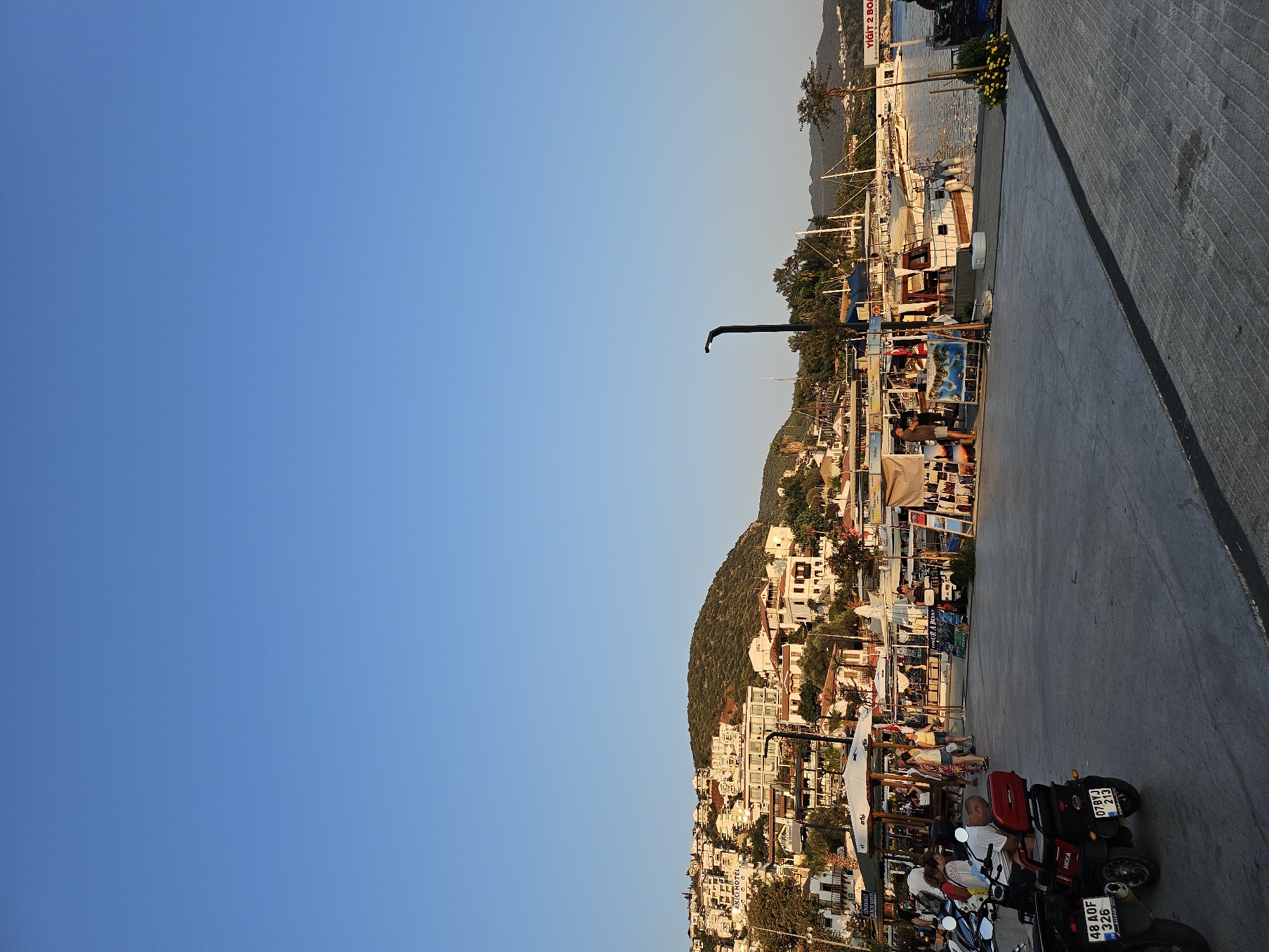 In an outdoor scene, a clear blue sky dominates the upper portion of the image. Below, a waterfront promenade bustles with activity. Buildings cascade up a hillside in the background, with white structures accented by terracotta roofs. Several boats are moored in the harbor. In the foreground, a paved area serves as a walkway and parking for several motorcycles and scooters. People are seen walking and standing around market stalls set up along the promenade, some displaying various goods. A lamppost stands near the water's edge.