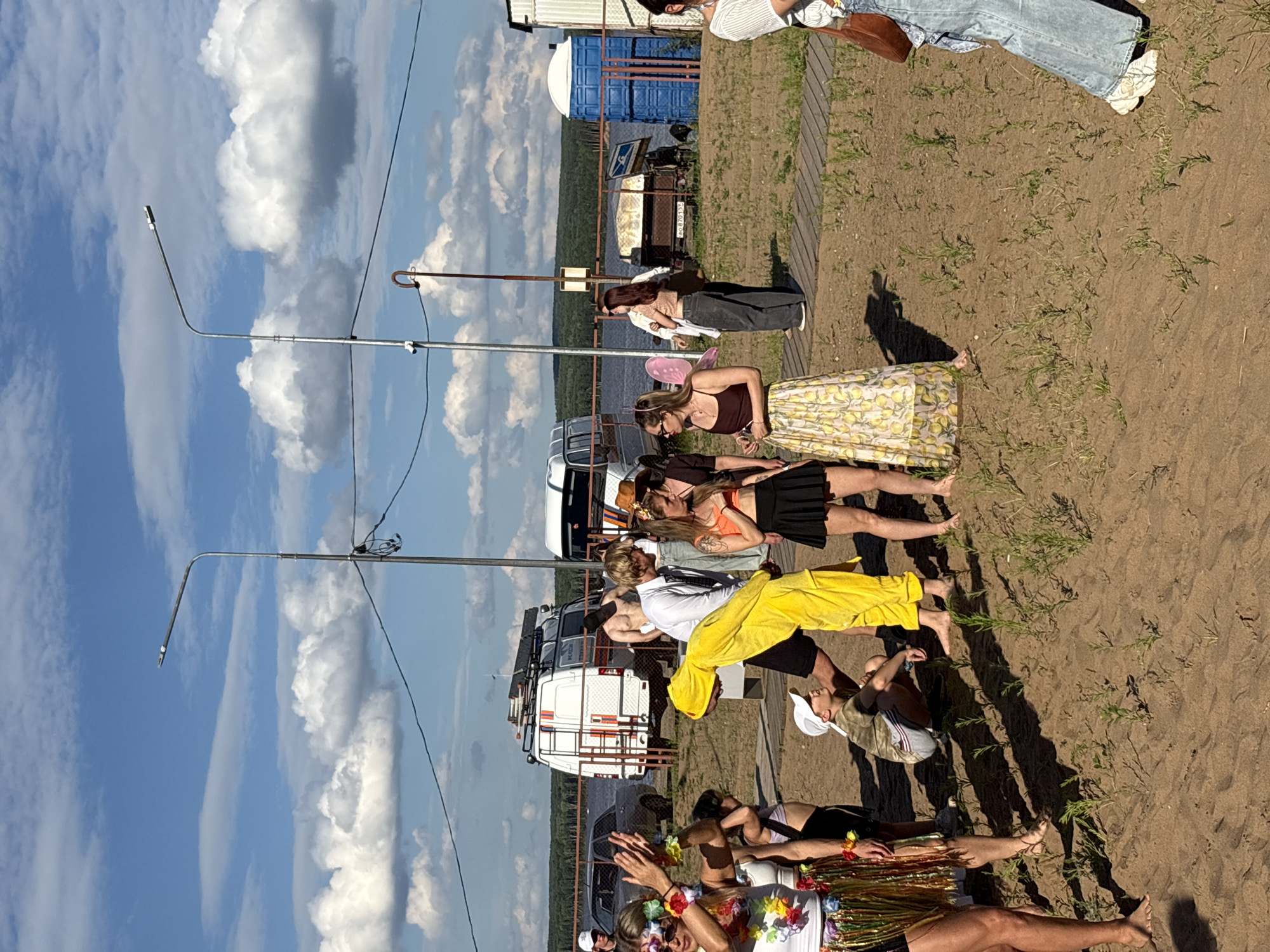 In the city of Chaikovsky, Russia, a group of people are gathered on a sandy area near a body of water. The sky is blue with fluffy white clouds. Two lampposts are visible in the background. Several vehicles are parked, including a white van with emergency markings and a smaller white van. A gray car is also present. In the foreground, several individuals are interacting. One person is wearing a bright yellow chicken costume. Another person is wearing a hula skirt and lei. Other individuals are dressed in casual clothing, including a woman in an orange top and black skirt, and a woman in a yellow skirt with a lemon pattern. There are portable toilets in the background. The ground is sandy with sparse vegetation.