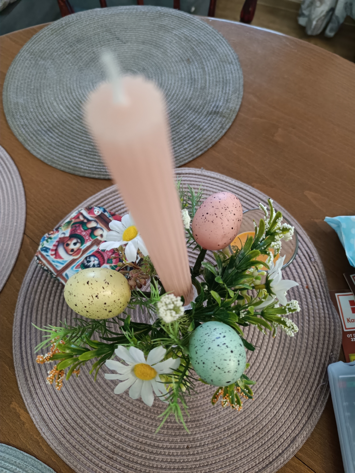 The image shows a centerpiece on a round, woven placemat atop a wooden table. The centerpiece features a thick, pale pink candle surrounded by artificial flowers and three speckled Easter eggs in yellow, pink, and blue. A small, colorful napkin with cartoon-like figures is visible near the yellow egg. Part of another placemat is visible on the table. In the background, there are parts of what appears to be a chair and curtains. The scene is likely inside a home. It is difficult to determine the location as Varna, Bulgaria, as there is no definitive evidence in the picture.