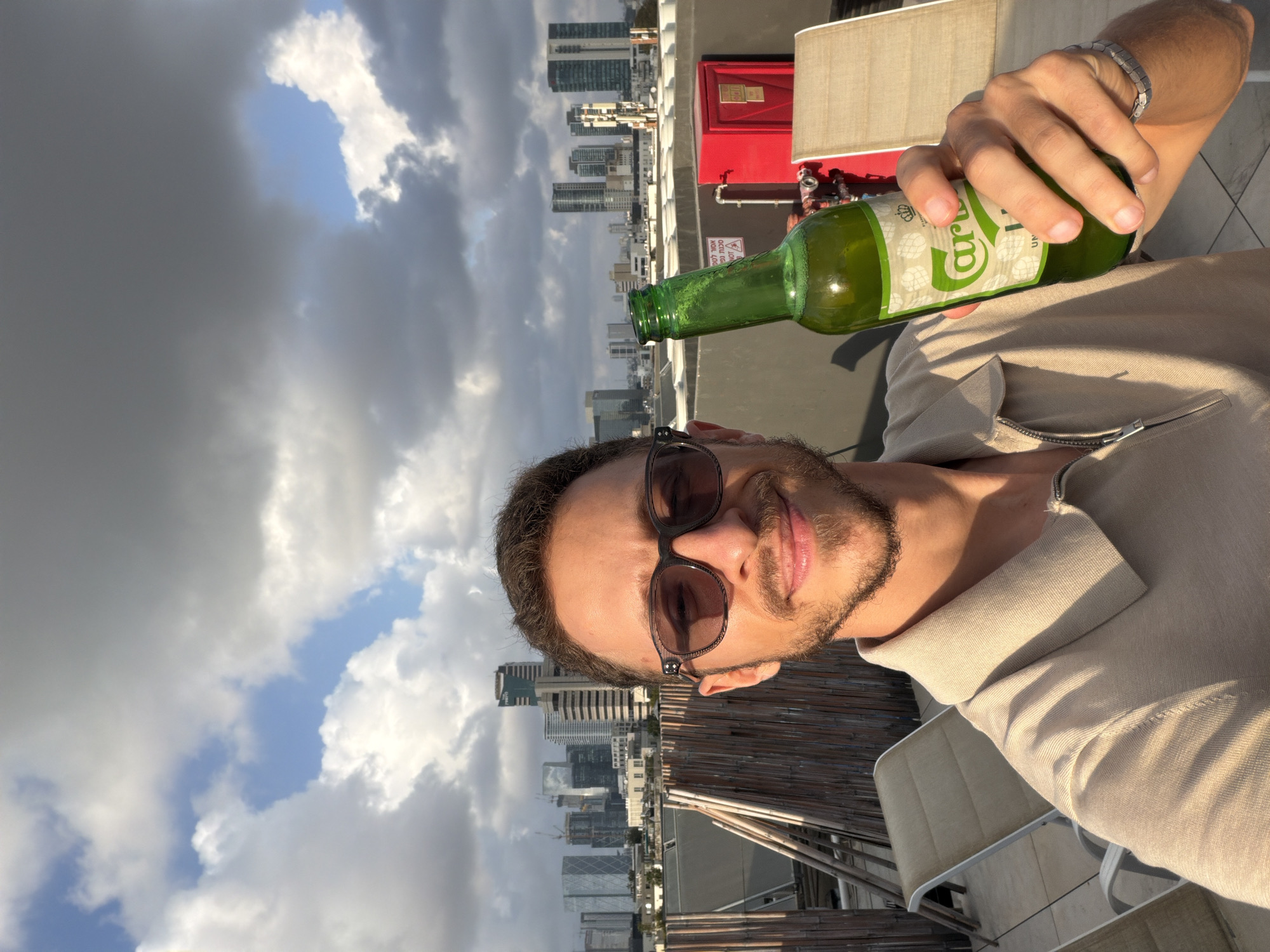 A man is on a rooftop, holding a green glass Carlsberg beer bottle. He is wearing sunglasses and a light tan collared shirt. A city skyline is in the background, indicating a city location. The skyscrapers suggest the location is Tel Aviv, Israel. The sky above features a mix of clouds and patches of blue sky. A red box and an outdoor lamp are visible in the midground. The man has a wristband on his wrist.