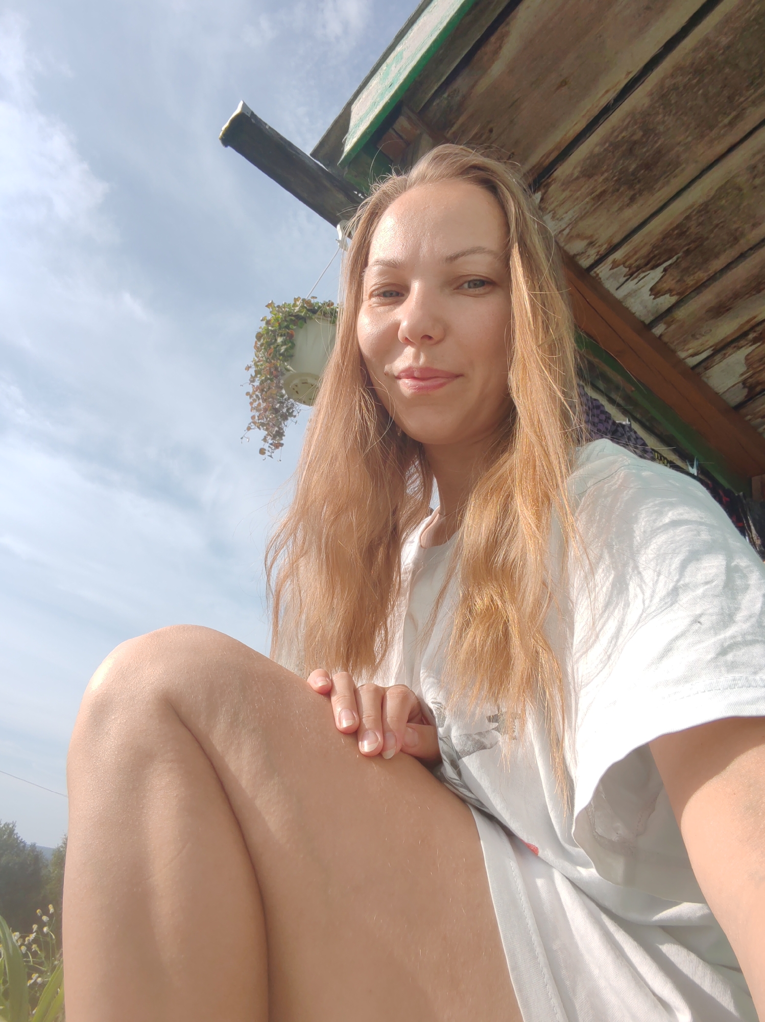 A close-up, low-angle selfie shows a young adult female with blonde hair, wearing a white t-shirt, sitting outdoors. She is positioned in the foreground, with her left leg bent and her right arm resting on it. Her gaze is directed towards the camera with a slight smile. Behind her, a rustic wooden structure with peeling paint and a green trim is visible overhead. A hanging plant in a white pot is suspended from the structure. The background consists of a bright blue sky with scattered white clouds, and a glimpse of greenery and trees can be seen on the left side of the image. The lighting suggests a sunny day. The location is specified as Чухлэм, Russia.