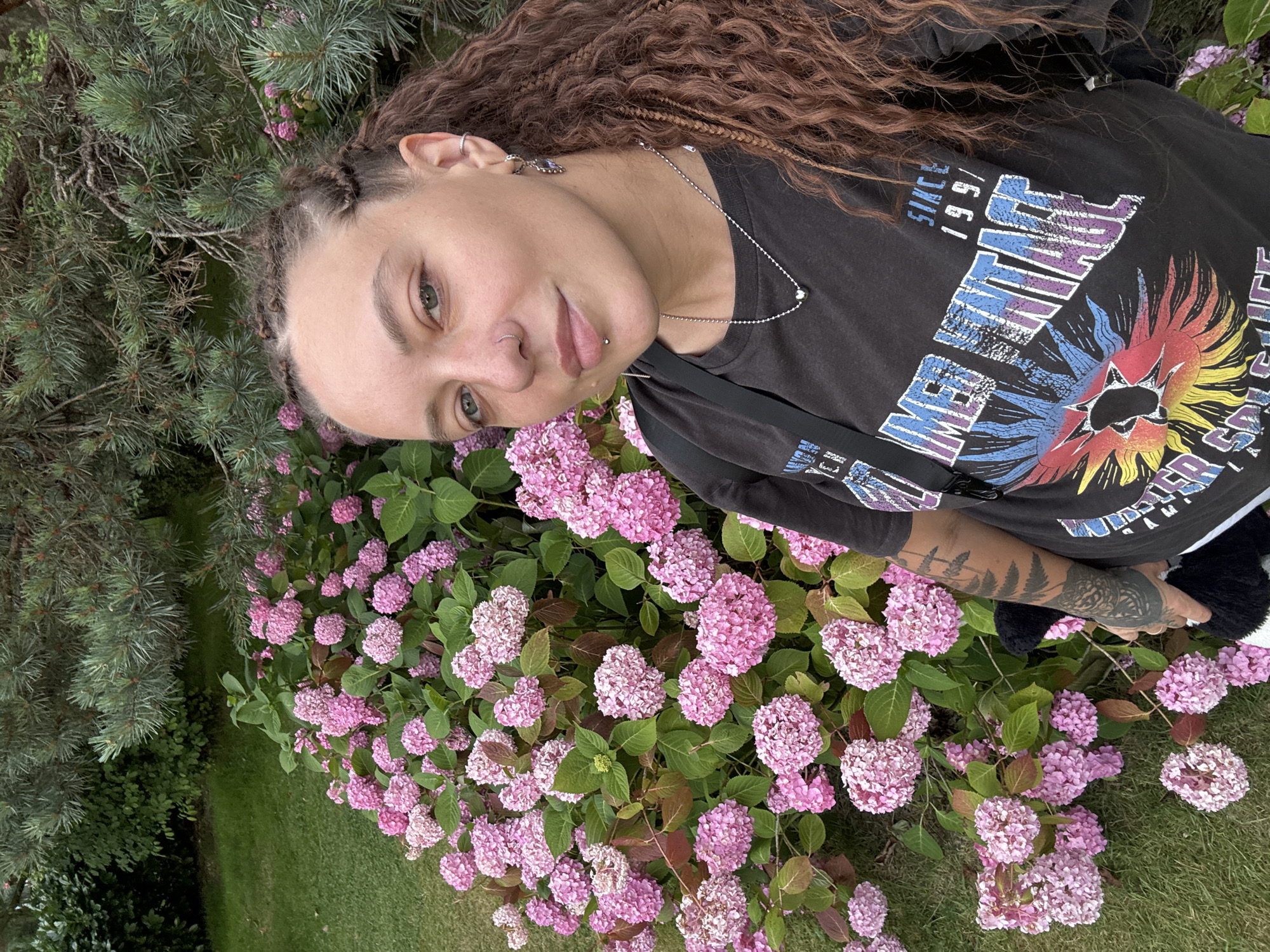 A young woman with braided hair and facial piercings takes a selfie in front of a bush of pink hydrangeas and a pine tree. She is wearing a black t-shirt with a graphic print and a silver necklace. The background consists of green grass and foliage. The woman's left arm is tattooed. There is no indication of the location being Sochi, Russia in the image.