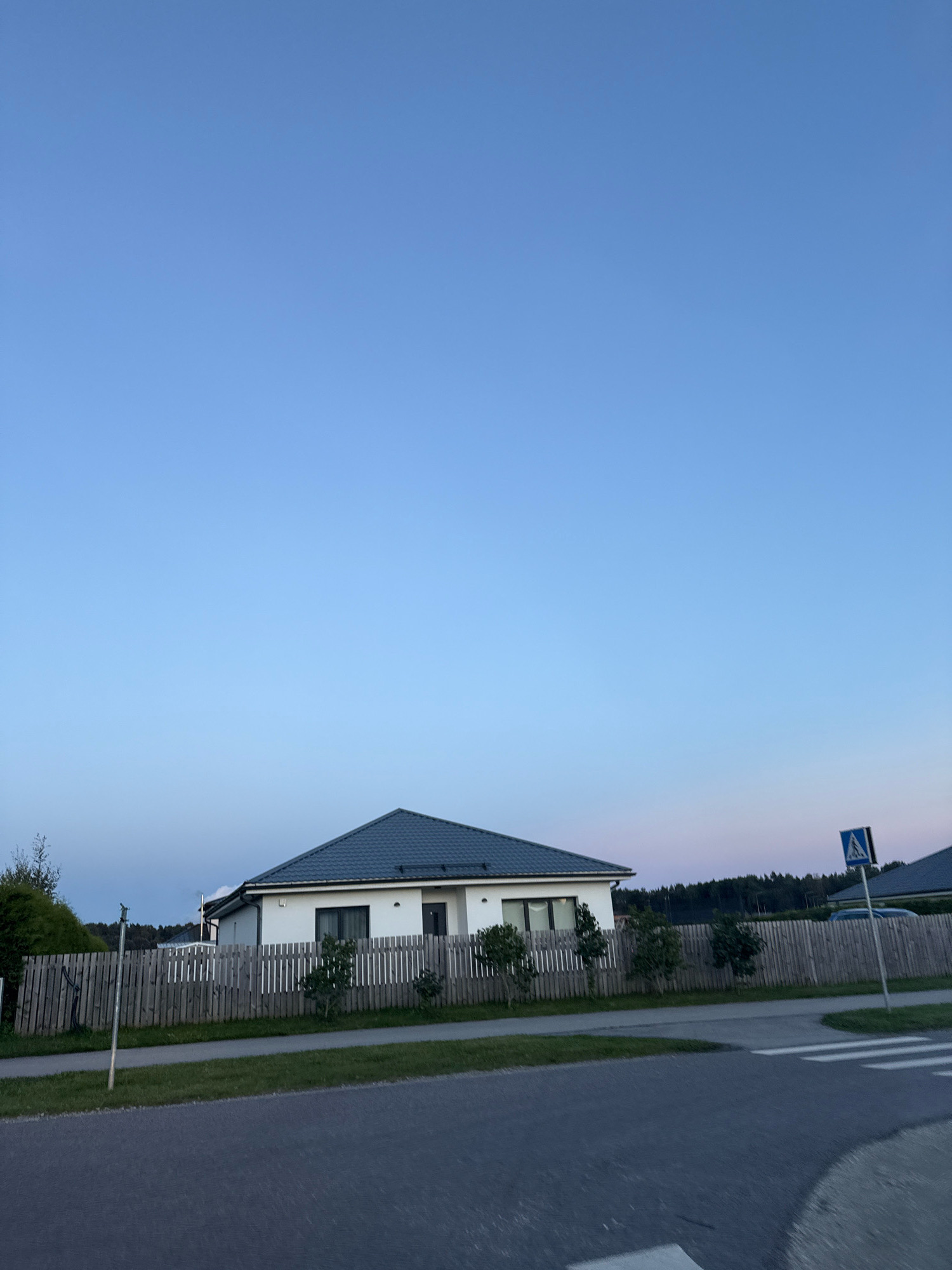 The image depicts a residential scene in Alliku, Estonia, under a clear blue sky transitioning to a soft pink and purple hue at the horizon. A modern, white, single-story house with a dark grey roof is the central focus, bordered by a weathered wooden fence. Small, manicured trees are planted in a row in front of the fence, separating the property from a paved road and a pedestrian crossing. A blue pedestrian crossing sign is visible on the right side of the image. The environment appears tranquil and suburban, with a line of trees forming the backdrop behind the houses.