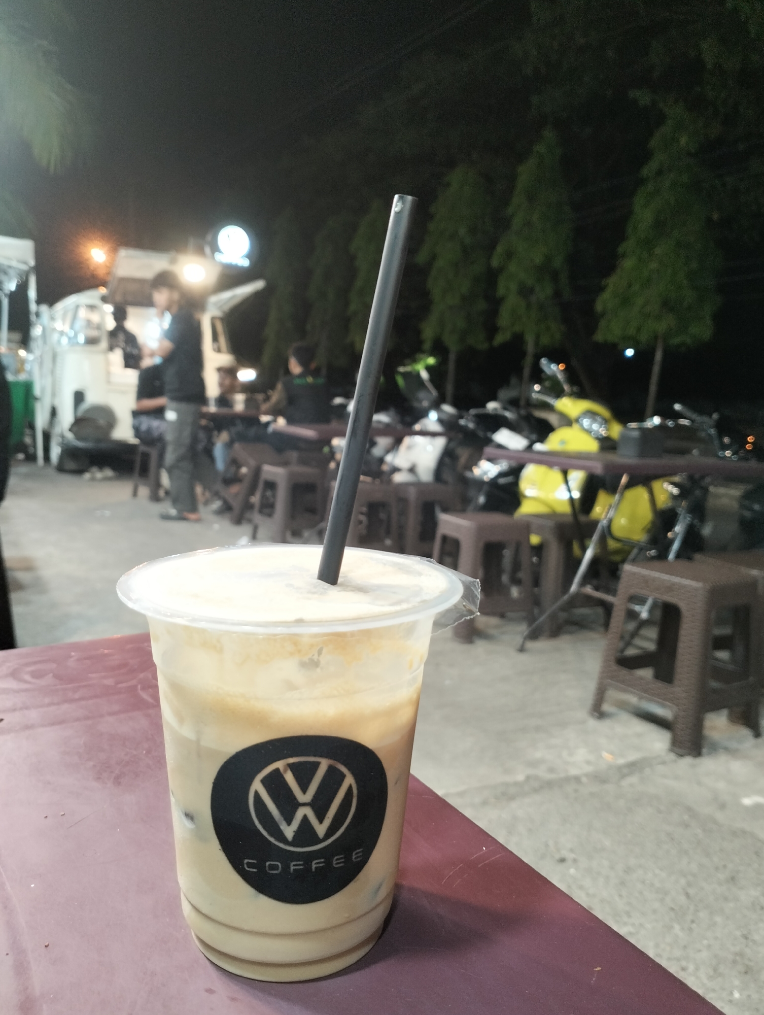 An iced coffee drink with a black straw and a "W Coffee" logo on the cup sits on a maroon table in the foreground. In the background, a food truck is visible, with several people standing and sitting around it. Motorcycles are parked in a line behind the food truck, and a row of trees lines the scene. The location appears to be an outdoor night market or street food area in Lhokseumawe, Indonesia.