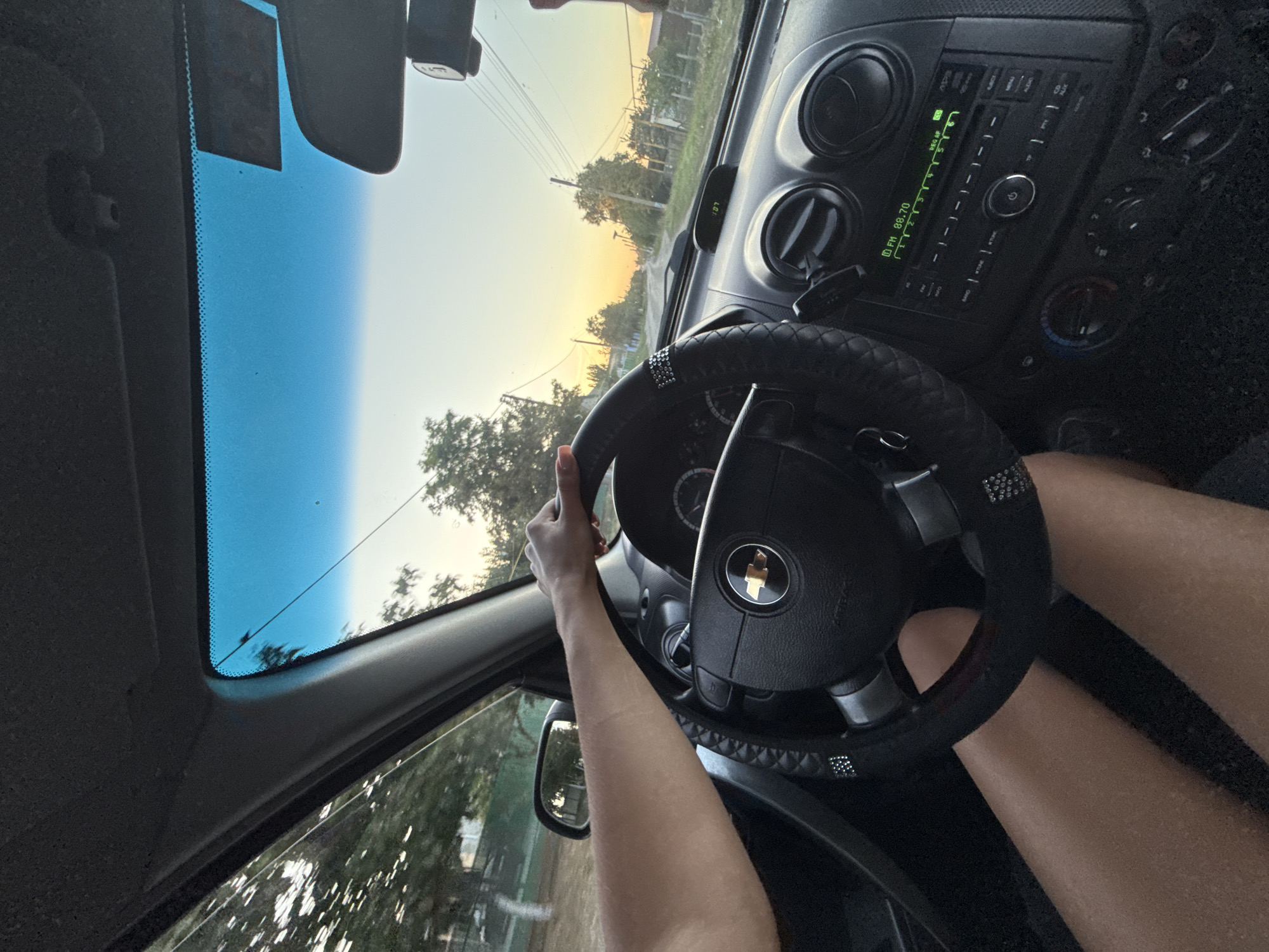 The image is taken from inside a car looking out through the windshield and side window. The driver's hands are on the steering wheel. The steering wheel is black and has a diamond-like trim. The dashboard of the car is visible, including the radio, air vents, and climate controls. The car is driving on a road in a rural area with trees and houses on either side. The sky is a clear blue with a hint of orange from the setting sun. The scene appears to be during the golden hour. Based on the provided context, the location is the city of Cioc-Maidan, Moldova.