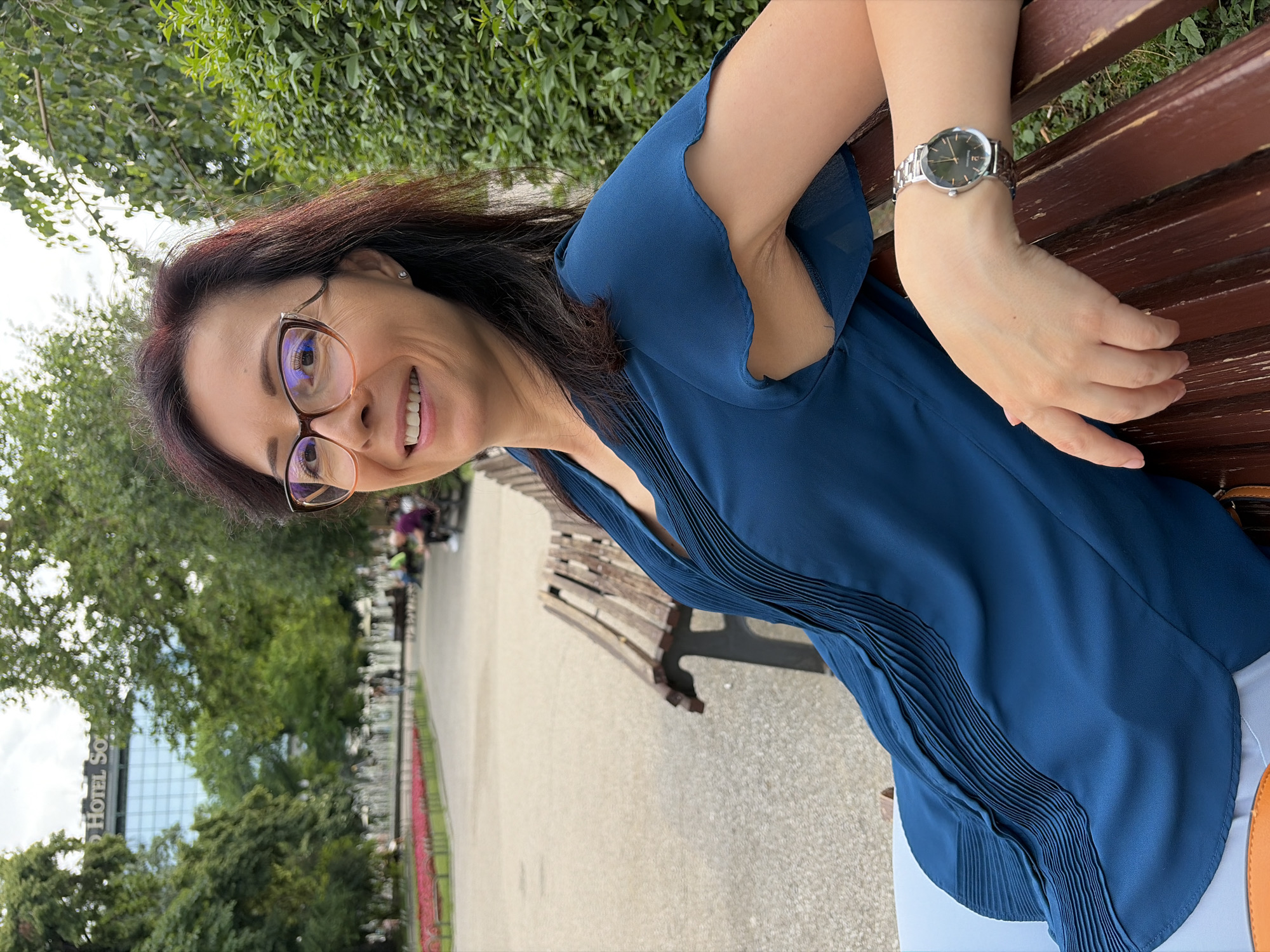 A woman with dark hair and glasses sits on a park bench in Sofia, Bulgaria. She wears a blue blouse and light blue pants. Her left arm rests on the bench, displaying a silver watch. In the background, a pathway leads past flower beds and a fountain, with a building labeled "HOTEL SOFIA" visible between the trees.