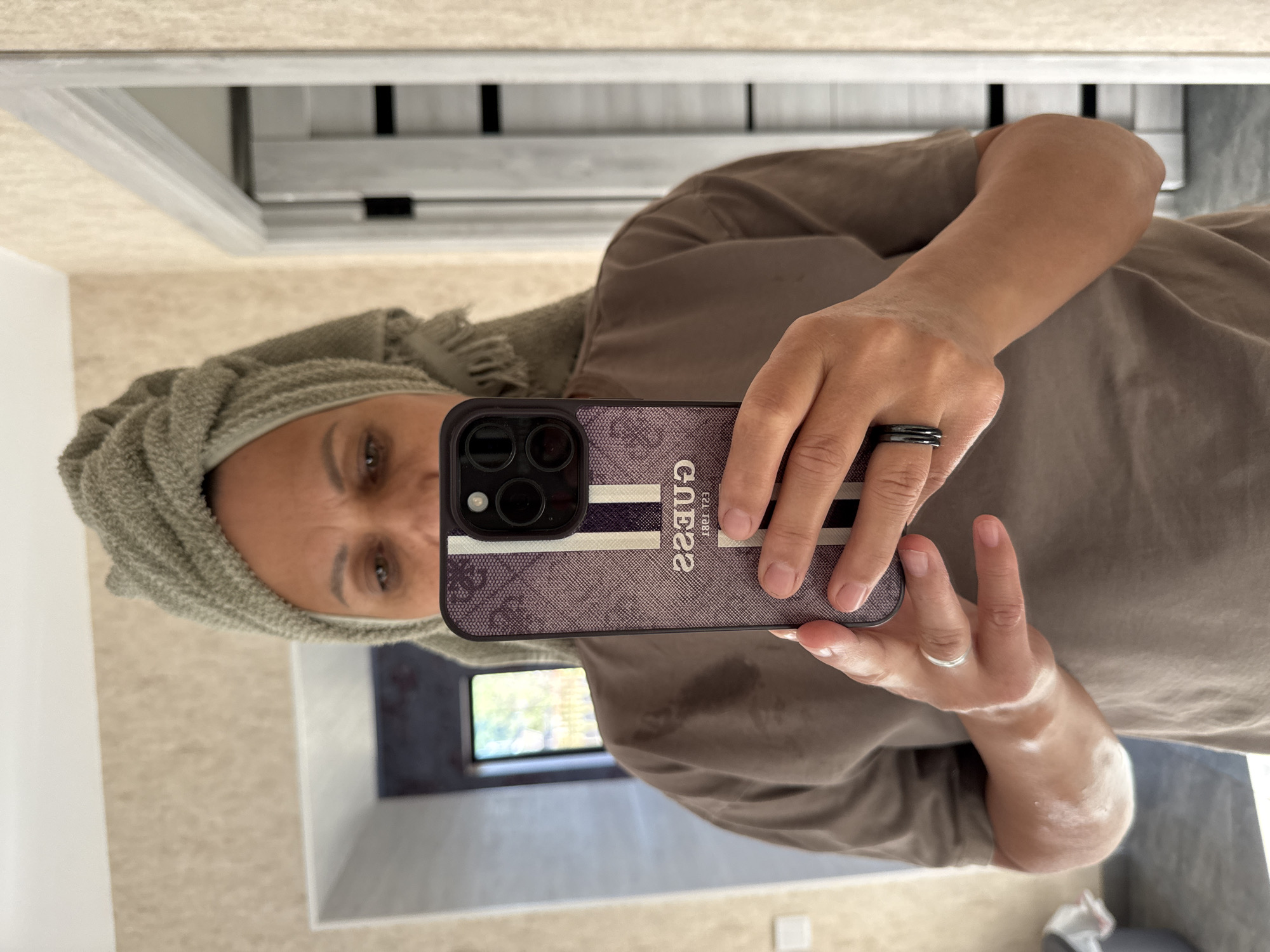A woman is taking a selfie in a bathroom. She has a towel wrapped around her head and is wearing a brown t-shirt. She is holding a smartphone with a purple and white striped case. A ring is visible on her left ring finger. The bathroom has beige textured wallpaper and a doorway with a white frame. The lighting suggests it is daytime. The location is not specified as Soloneshnoye, Russia in the image.