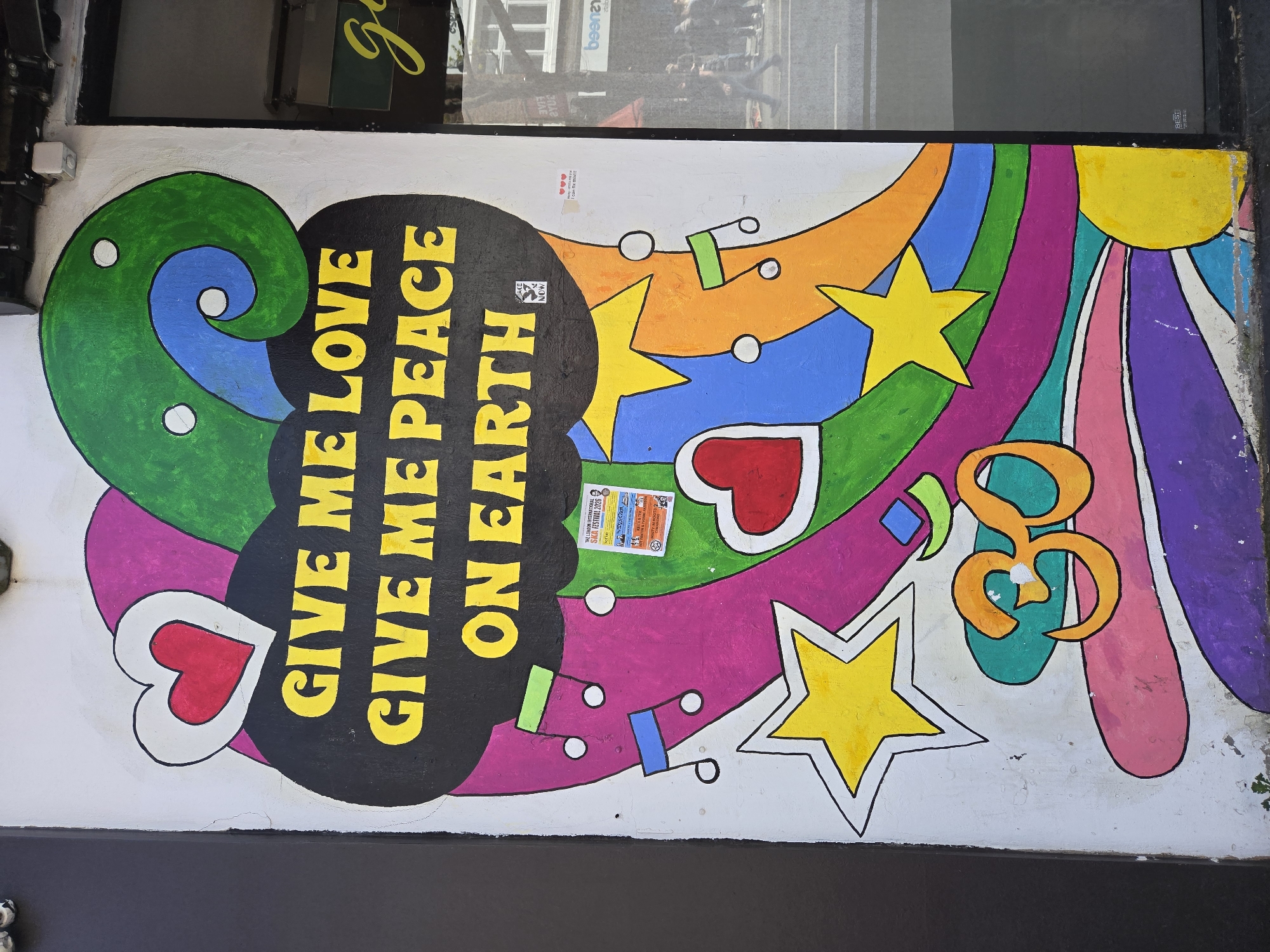 Here is a description of the image:
This is a photograph of a colorful mural on a white wall. The mural features a psychedelic design with swirling shapes, stars, hearts, and musical notes in bright colors like green, blue, orange, yellow, pink, and purple. The text "GIVE ME LOVE, GIVE ME PEACE ON EARTH" is painted in yellow letters inside a black, cloud-like shape.
A sticker and a poster are pasted on top of the mural. The poster refers to "THE LONDON INTERNATIONAL SIKA FESTIVAL 2026" and has additional text in smaller print.
The mural appears to be located on the exterior of a building, possibly a shopfront, as indicated by the dark-colored structure at the bottom of the frame. The location is in the city of London, United Kingdom based on the wording of the poster. The mural has a playful and peace-oriented theme.