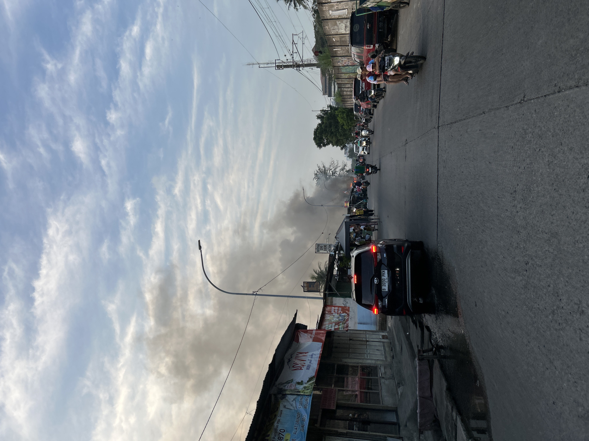 The image depicts a street scene in an urban environment, possibly a market area, with a significant amount of dark smoke billowing into the sky from the left side of the frame. The smoke appears to be originating from a fire. The street is paved and lined with buildings and stalls on the left, and a wall with some vegetation on the right. A variety of vehicles are present on the road, including cars and numerous motorcycles, some carrying multiple passengers. People are visible walking and riding on motorcycles. The sky is partly cloudy with daylight illumination. The overall atmosphere suggests a bustling street with an ongoing event causing heavy smoke.
