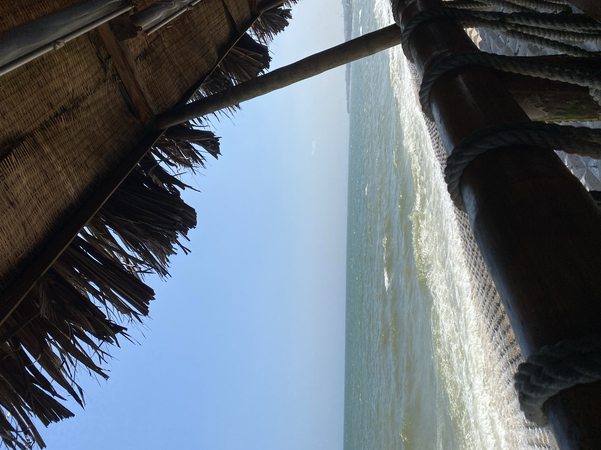 A view of the ocean from beneath a thatched roof structure. The roof, made of dried palm fronds, overhangs the scene, with some fronds hanging down. A thick wooden beam, wrapped with thick rope, runs horizontally across the foreground, obscuring the lower portion of the beach and shore. Beyond the beam, the ocean stretches out under a clear blue sky, with moderate waves breaking on a stony shore. In the distance, a faint coastline is visible, suggesting the location is Mũi Né, Vietnam. The scene is devoid of people or any obvious activity.