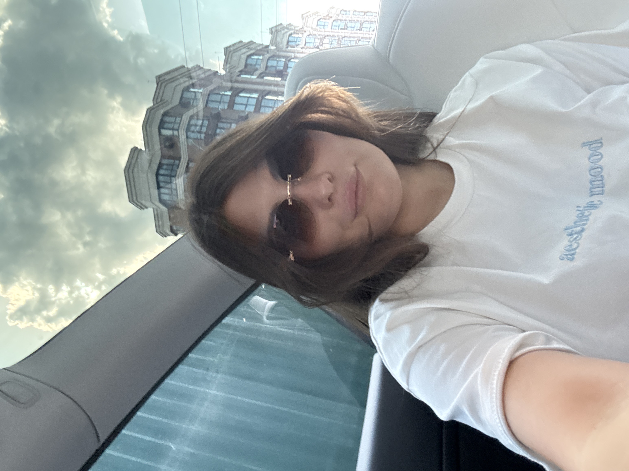 A young woman takes a selfie inside a car, captured at a tilted angle. She is in the foreground, facing the camera with a neutral to slight smile, wearing dark-tinted, gold-framed sunglasses. Her medium-length dark brown hair partially frames her face. She wears a plain white short-sleeved t-shirt with light blue text on the chest that reads "aesthetic mood."
The setting is inside a vehicle, evidenced by the light-colored car seat behind her and parts of the car's interior, including a window frame and roof lining. Through the car window, a large, multi-story building with a light-colored, detailed facade and many windows is visible in the background. The sky beyond the building is cloudy, with varying shades of grey, indicating daytime. Reflections of the sky and a distorted view of the building are present on the car's windows. The overall mood is casual and relaxed. The location context for this image is Sofiivska Borshchahivka, Ukraine.