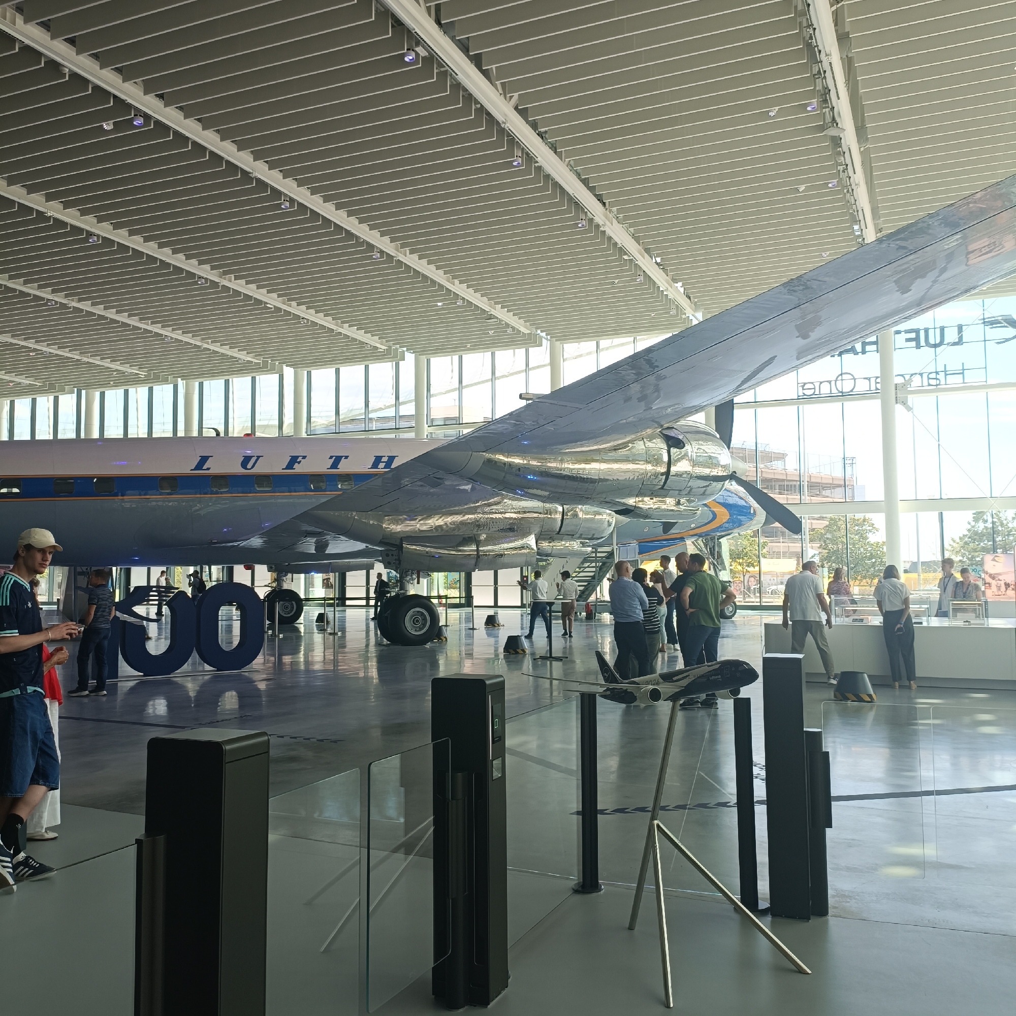 The image depicts the interior of a museum exhibit hall in Frankfurt, Germany, featuring a vintage Lufthansa airplane. The large aircraft, with its polished metal fuselage and prominent "LUFTHANSA" text, is the central subject. In the foreground, large, dark blue, three-dimensional numerals "100" are displayed, likely celebrating a centenary. Several people are present, observing the exhibits. Some are standing in groups, while others are walking. A man in a blue shirt and shorts stands on the left, looking towards the airplane. The environment is a modern building with a high, slatted ceiling, large windows offering views of the exterior, and a polished concrete floor. A small model airplane on a stand is visible in the lower right corner, along with exhibit barriers and display cases. The lighting suggests an indoor daytime setting.