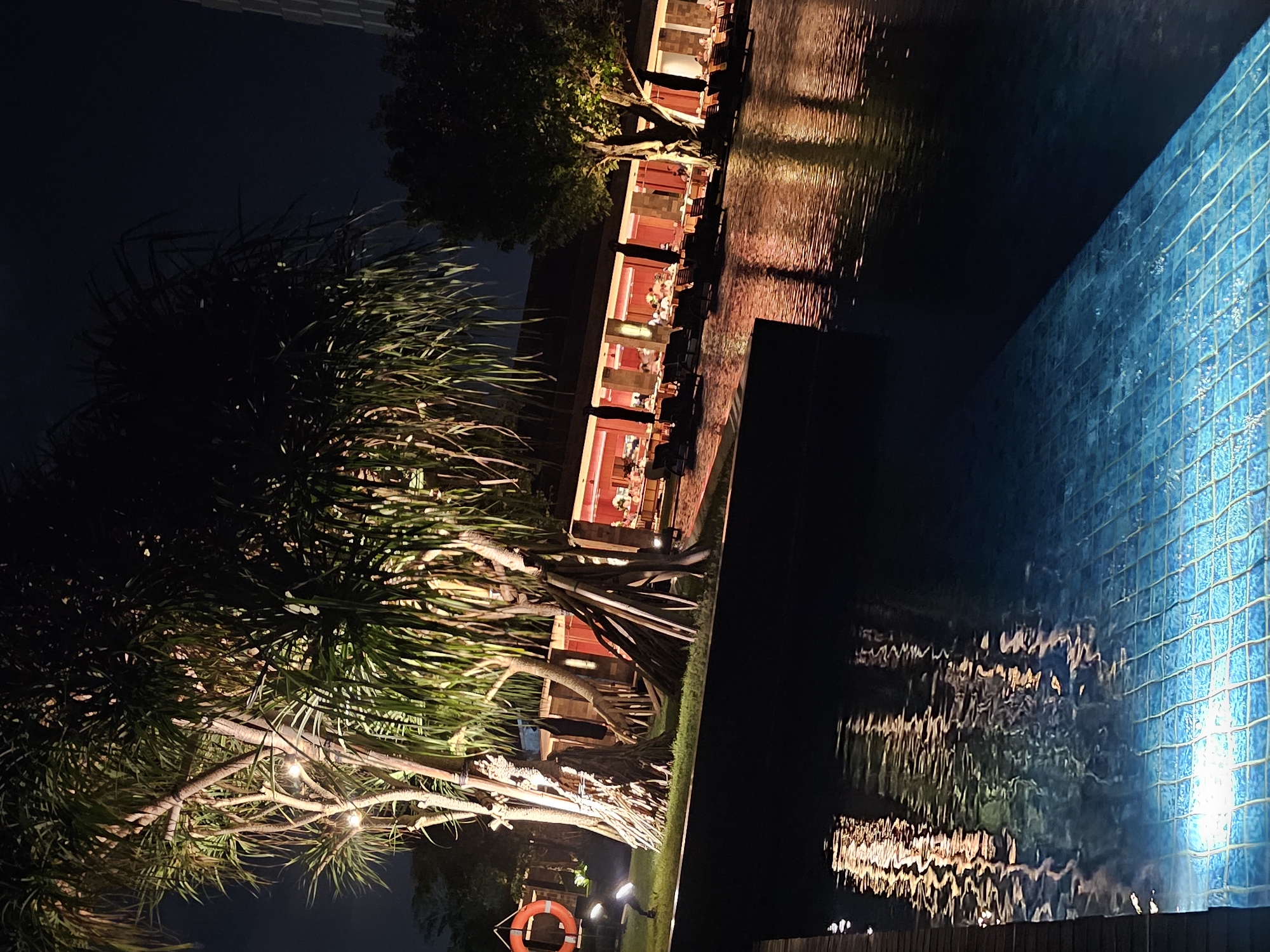 This is a nighttime exterior shot of a resort in Denpasar, Indonesia, featuring a swimming pool in the foreground and a restaurant in the background. The pool is illuminated with blue underwater lights, creating a shimmering effect on its tiled surface. Reflections of the warm lighting from the restaurant and surrounding foliage are visible on the water's surface.
To the left, a lifesaver ring is mounted on a post, and a floodlight is positioned on the ground. Several trees with distinctive, thick trunks and spiky fronds, characteristic of pandanus trees, are lit from below, casting dramatic shadows. They are supported by diagonal wooden braces.
The restaurant, a long, low building with an open-air seating area, is brightly lit with warm orange and red tones. Tables and chairs are set up, and several patrons are visible dining or socializing. Umbrellas are placed over some of the tables.
In the background, to the right, a portion of a modern building with a light-colored, textured facade is visible against the dark night sky. The overall atmosphere is one of serene, upscale evening dining and relaxation.