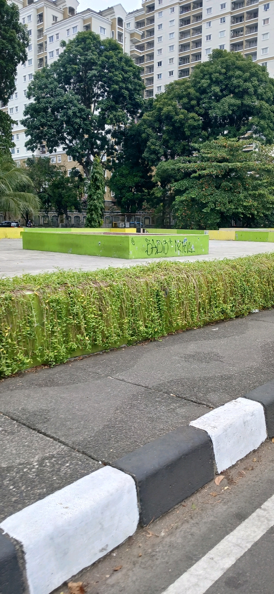 The image shows a street scene in North Jakarta, Indonesia, with a residential building and trees in the background. In the foreground, there is a curb with alternating black and white painted sections, separating the asphalt road from a concrete sidewalk. A low green wall with hanging vines runs along the edge of the sidewalk. Behind this wall, there is a bright green rectangular structure with graffiti on it, possibly part of a park or recreational area. A few trees are visible, and the sky is overcast.