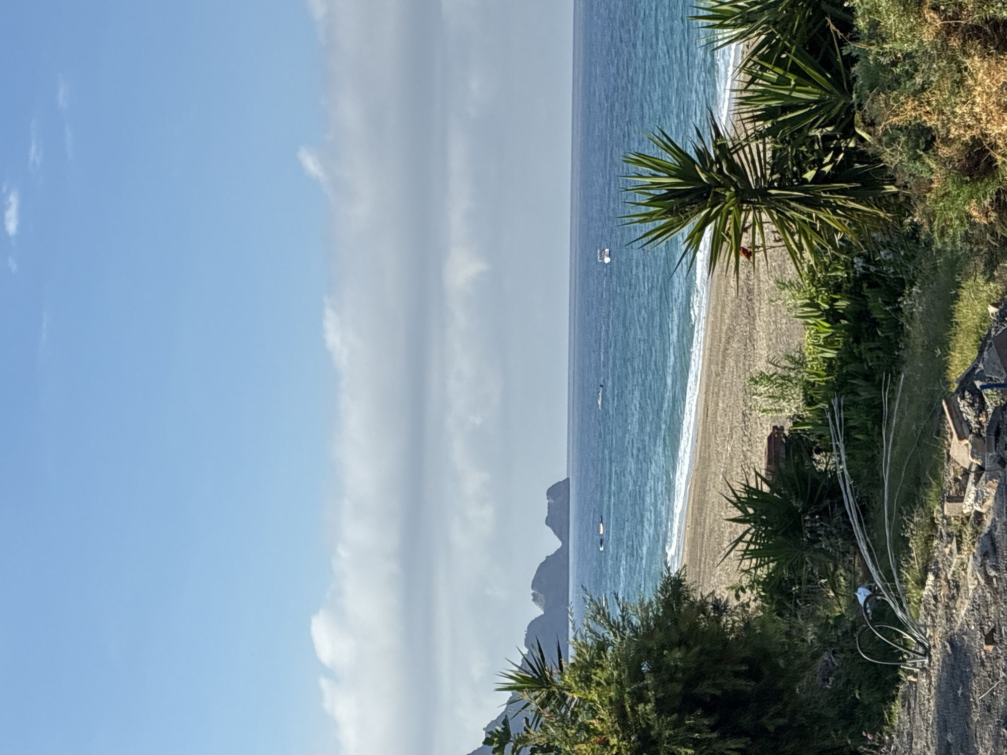 The image depicts a coastal scene in Alanya, Turkey, on a clear day with a bright blue sky and scattered clouds. In the foreground, there is a dirt path with some debris, including bricks and coiled cables. Lush green vegetation and palm-like plants are visible on the right and left sides, partially framing the view. The middle ground features a sandy beach with gentle waves lapping at the shore. Beyond the beach, the deep blue Mediterranean Sea stretches to the horizon. In the distance, rocky islands or promontories jut out from the water. Several small boats are scattered on the sea. The overall impression is of a serene and natural coastal landscape.
