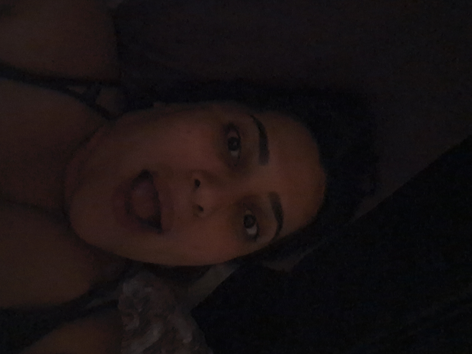 The image is a close-up, horizontally oriented, low-light selfie taken from a slightly upward angle. It features a person lying down, with their face taking up a significant portion of the frame. The person's eyes are open and looking towards the camera. Their mouth is slightly open. The lighting is dim, creating a dark and somewhat blurry overall image. The background is also dark and indistinct, with a hint of fabric visible to the upper right. There are no identifiable landmarks or contextual clues within the image to suggest a specific location in Manisa, Turkey.