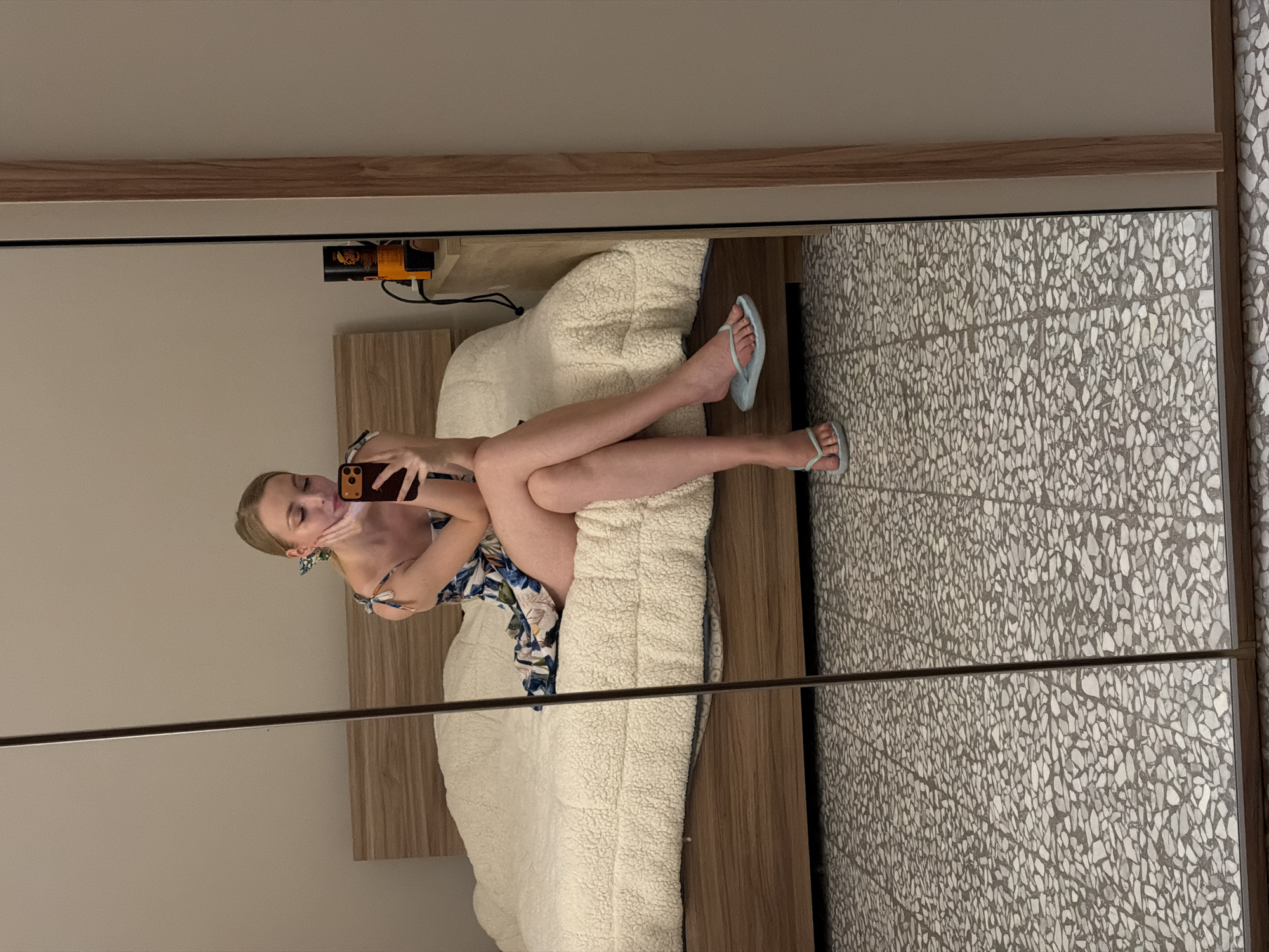 A woman is reflected in a mirror while sitting on a bed. The room appears to have a light-colored wall, a patterned floor, and a wooden frame around the mirror. The woman is holding a mobile phone and seems to be taking a selfie. She wears a patterned dress and light blue flip-flops. There is a bedside table with items on it near the bed. Without additional information, it's not possible to pinpoint the exact location within Catania, Italy.