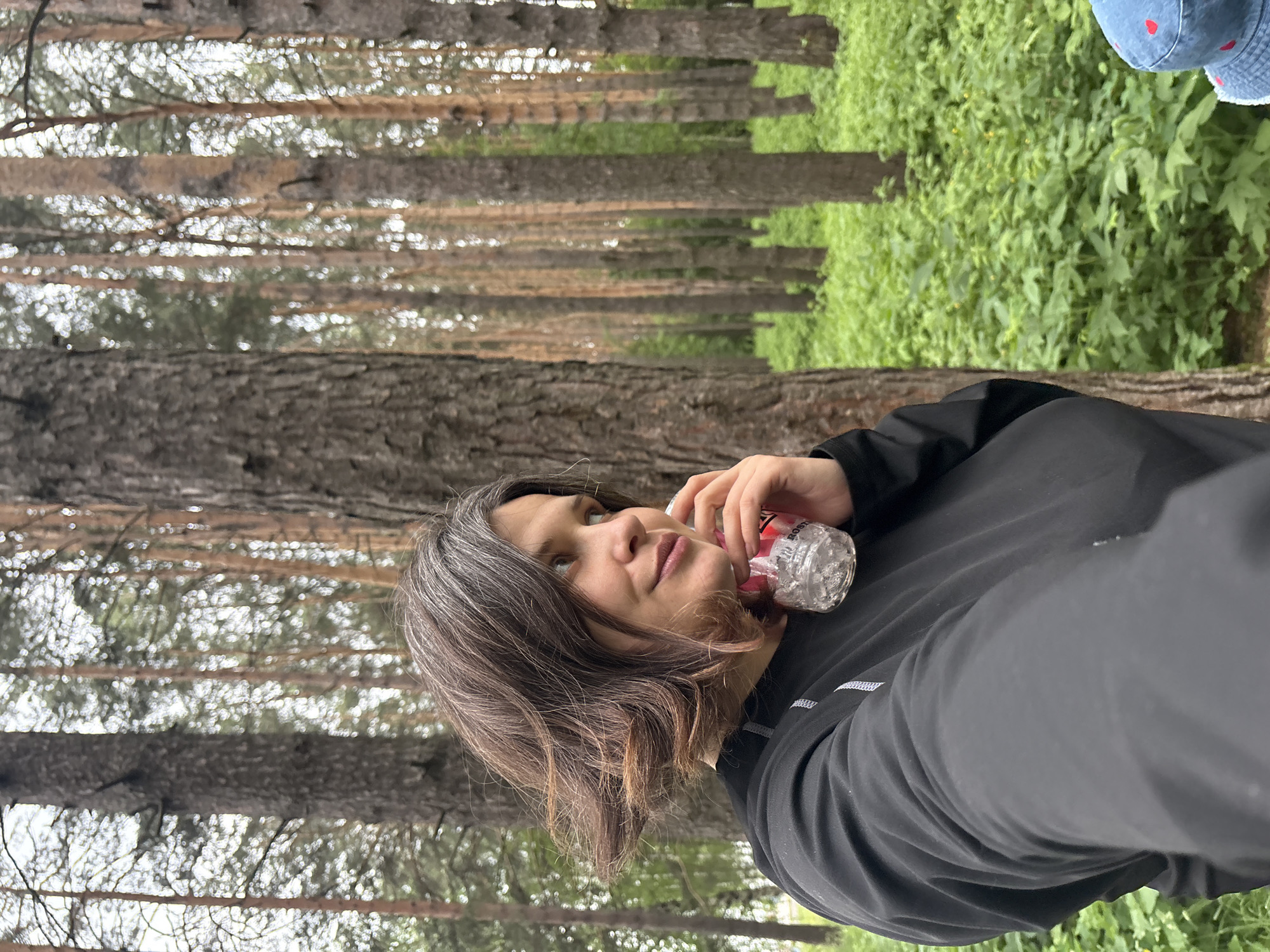 A woman with short, layered brown hair with gray streaks is in a forest. She is wearing a black long-sleeved shirt. She is holding a clear plastic bottle to her right ear, appearing as if she is on the phone. She is looking to her right with a slight smile. The background is a dense forest with tall pine trees and green foliage. The lighting is overcast.