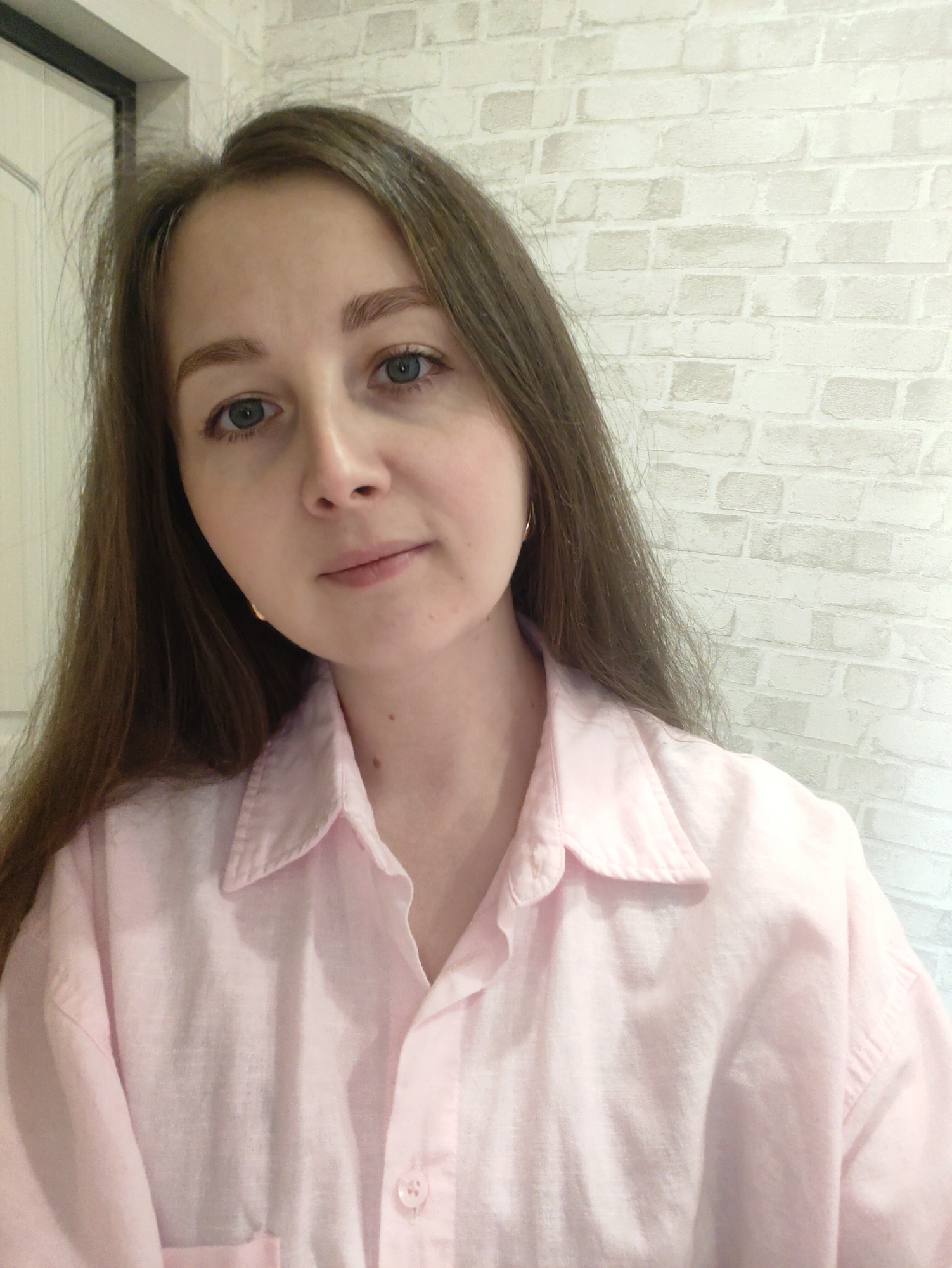 A head-and-shoulders portrait features a woman with fair skin, long, light-brown hair, and blue eyes looking directly at the camera. Her expression is neutral, with a very subtle hint of a smile. Her hair is parted to the left and appears slightly dishevelled or voluminous around the crown. She is wearing a light pink, collared shirt, likely made of linen or cotton, with the top buttons undone. A small, golden-colored hoop earring is visible on her left ear.
The background primarily consists of a light-colored wall with a brick pattern. To the far left, a vertical section of a white door or door frame is visible. The lighting is soft and even, suggesting an indoor setting. There is no visible text in the image. The overall scene is a casual, close-up portrait.