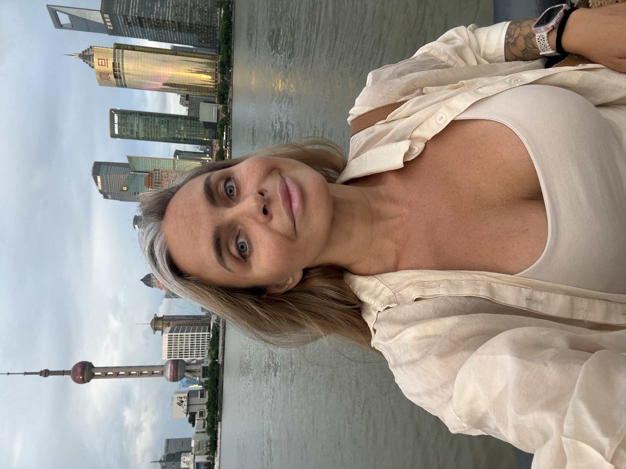 A woman with blonde hair and blue eyes takes a selfie on a waterfront with the Shanghai skyline in the background. She is wearing a white tank top and a loose, light-colored button-down shirt. Her left arm is resting on her hip, and her right arm is extended to hold the camera. The Shanghai skyline features several modern skyscrapers, including the Oriental Pearl TV Tower, and is reflected in the water. The sky is overcast.