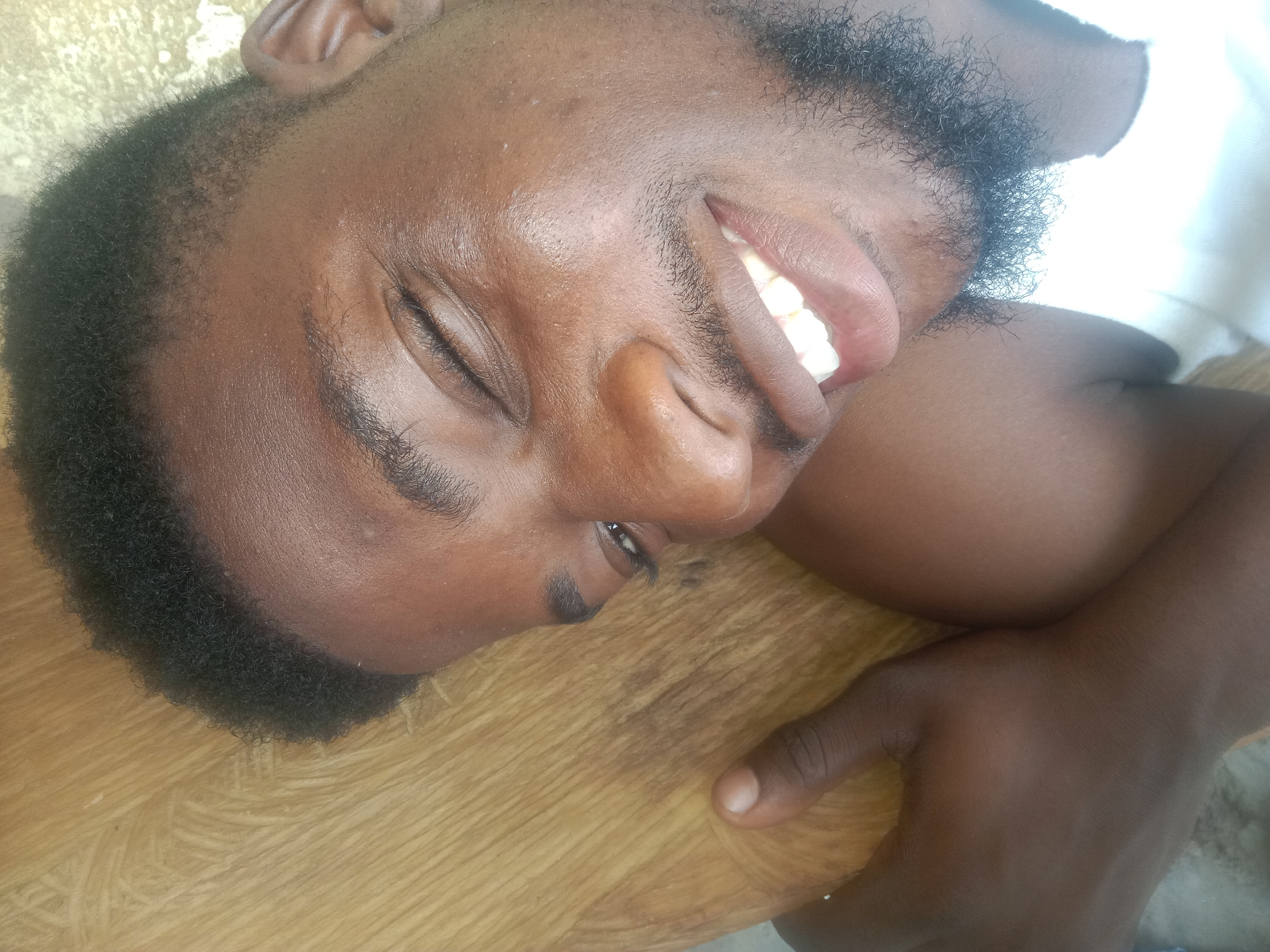 A close-up portrait of a person with dark skin, a beard, and dark hair is shown, lying on a light brown wooden surface. The person's eyes are mostly closed, and they are smiling, revealing their teeth. The location and city are unknown.