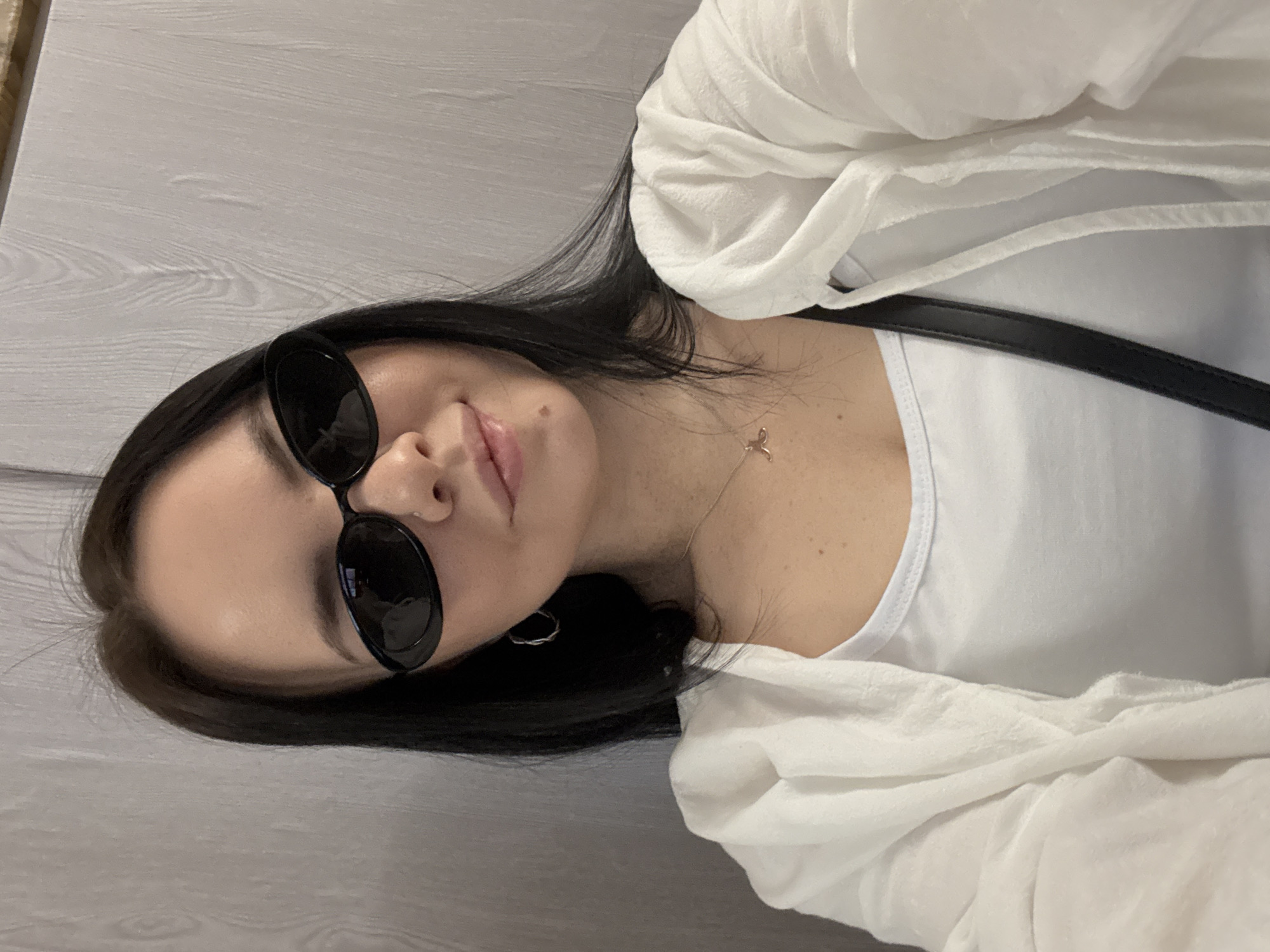 A young woman with dark hair is posing for a selfie. She is wearing a white t-shirt and a white, loose-fitting overshirt. She has dark sunglasses on and a necklace with a small pendant. A black purse strap is visible across her chest. The background appears to be a light-colored wall or surface with a wood-grain pattern. The location is not identifiable as Mytishchi, Russia based on the provided image.
