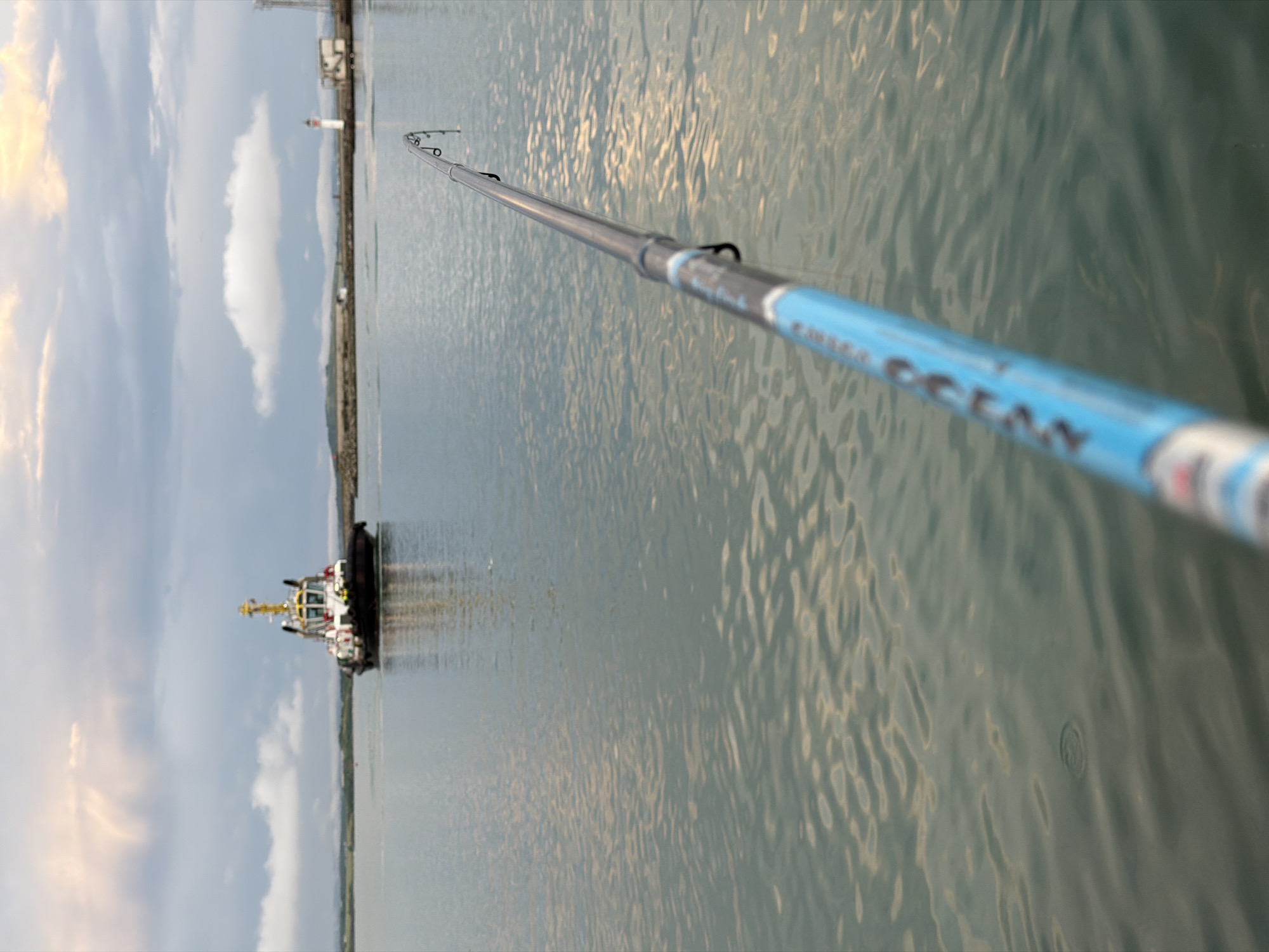 The image captures a serene coastal scene from a low-angle perspective, centered on a fishing rod extending diagonally across the frame. The rod, grey with a distinctive bright blue section closer to the viewer, stretches from the lower right towards the upper left of the image, appearing to point towards the distant horizon. On the blue part of the rod, the text "OCEAN" is clearly visible, suggesting a brand name, with other letters possibly obscured.
The majority of the background and midground is dominated by the calm, rippling surface of the water, reflecting the soft, warm light of what appears to be late afternoon or early evening. In the mid-distance, on the left side of the frame, a white and yellow tugboat floats on the water. Further along the horizon, a pier or breakwater is visible, with a small white lighthouse marked with a red stripe. The sky above is partly cloudy, with some clouds illuminated by the setting sun, adding to the tranquil atmosphere.
The primary activity suggested is fishing, with the rod positioned over the water. No people are visible in the frame. The scene provides a glimpse into a peaceful moment in a harbor or coastal environment, consistent with the provided context of Burgas, Bulgaria.