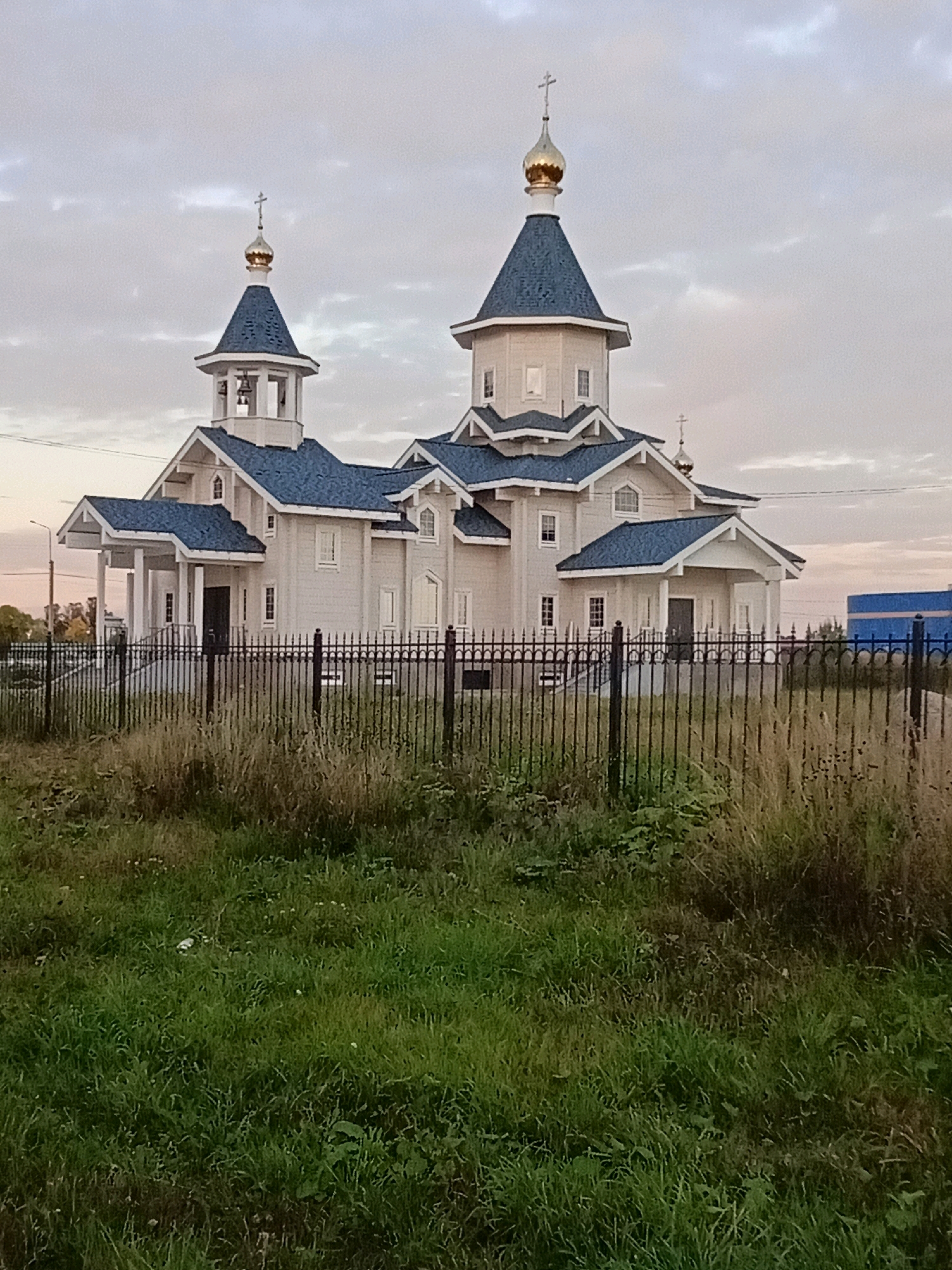 A light-colored, multi-tiered wooden church with blue roofs and gold domes stands behind a black metal fence. A bell tower, also with a blue roof and gold dome, is to the left of the main structure. The church has several windows and an entrance with a porch. The foreground is a grassy field with tall weeds. In the background, a blue building and telephone lines are visible under a cloudy sky.