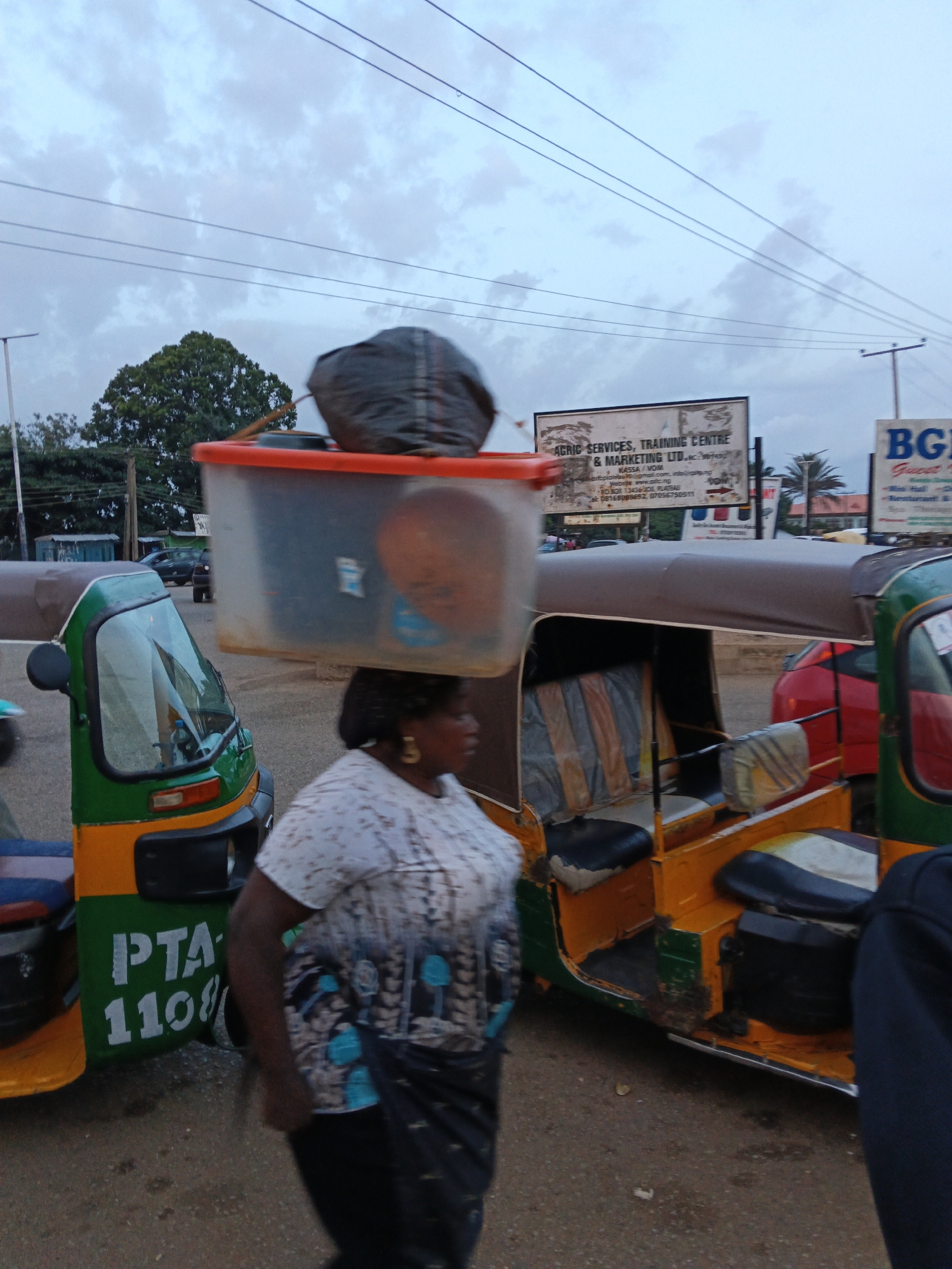 A woman carries a large clear plastic tub filled with items and a dark bag on her head as she walks past a yellow and green auto-rickshaw. The woman is wearing a white t-shirt with a patterned design and a dark skirt. The auto-rickshaw has the text "PTA 1108" printed on its side. In the background, there are more auto-rickshaws, a red car, trees, utility poles, power lines, and billboards. One billboard reads "AGRIC SERVICES, TRAINING CENTRE & MARKETING LTD.". The sky is overcast.