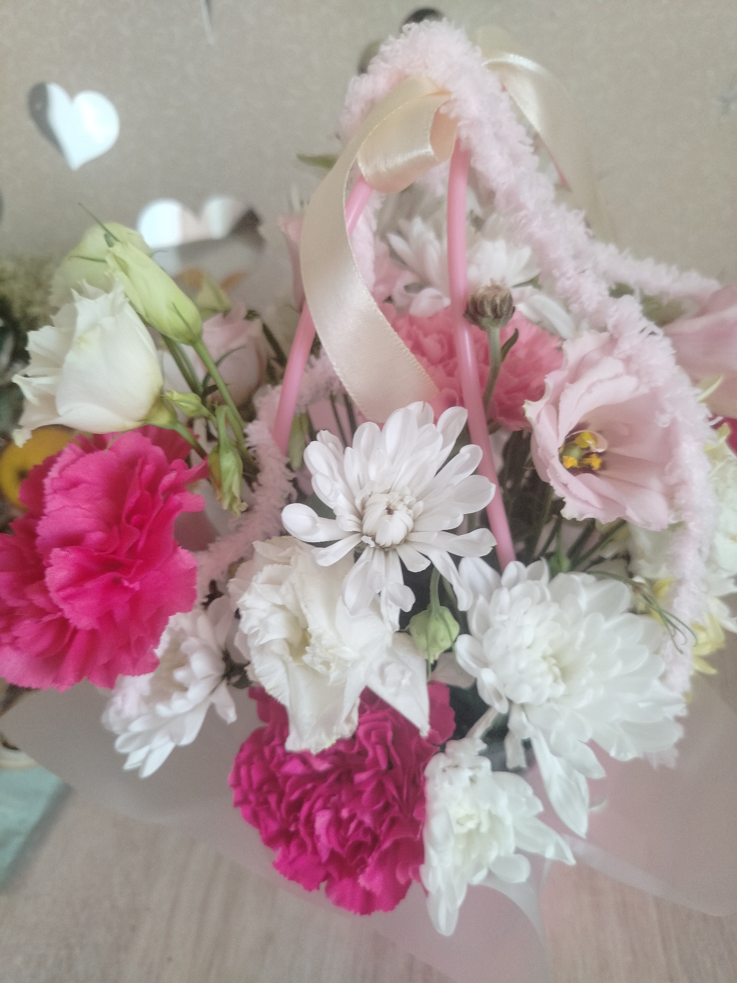 A close-up, eye-level shot captures a colorful floral arrangement. The bouquet is presented in a pink basket, adorned with a light pink fuzzy trim and a pale peach ribbon. The flowers themselves are a mix of vibrant pink carnations, delicate white chrysanthemums, and soft pink lisianthus. Green buds and stems are also visible. The background is a textured, light-colored wall with subtle heart-shaped cutouts, one of which is illuminated. The surface below the arrangement appears to be a light-colored, wood-like material. The scene appears to be indoors, with soft lighting. The city of Yashkino, Russia is not depicted in this image.
