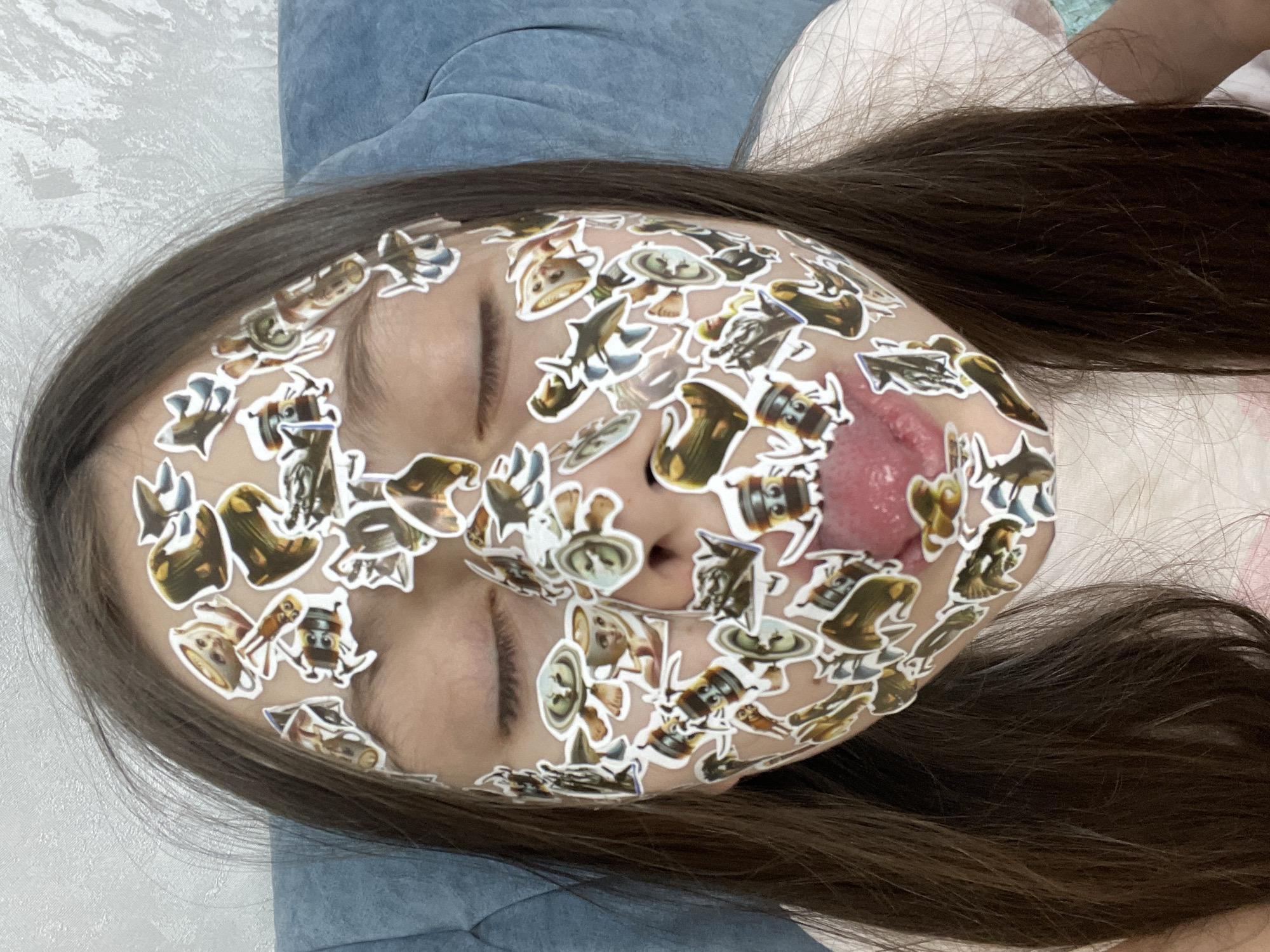 A young individual is lying down, with stickers covering their face. Their eyes are closed, and their tongue is slightly out. The background includes bedding and a blue cushion. No information indicates the location is Izhevsk, Russia.