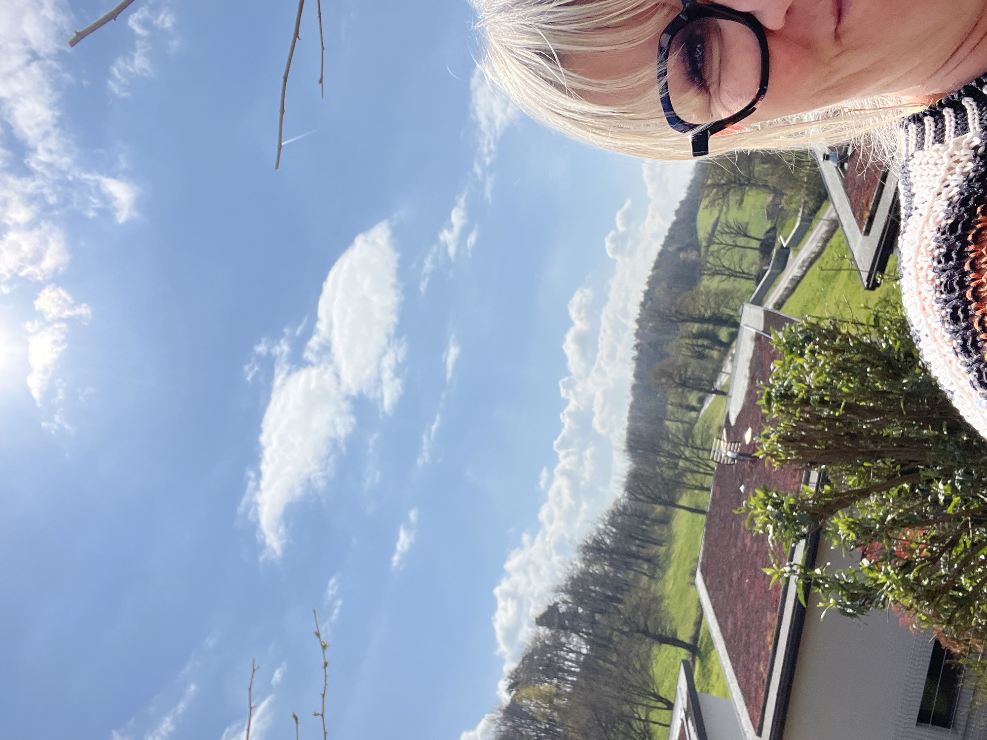 A daytime outdoor photo, likely a selfie, features a partial view of a person with blonde hair and glasses in the upper right corner. The background reveals a landscape with trees, a green field, and rooftops of buildings. The sky is blue with scattered clouds. The location is likely Chambœuf, France. The photo's orientation is slightly rotated counter-clockwise. A tree branch is visible in the lower right and upper left.