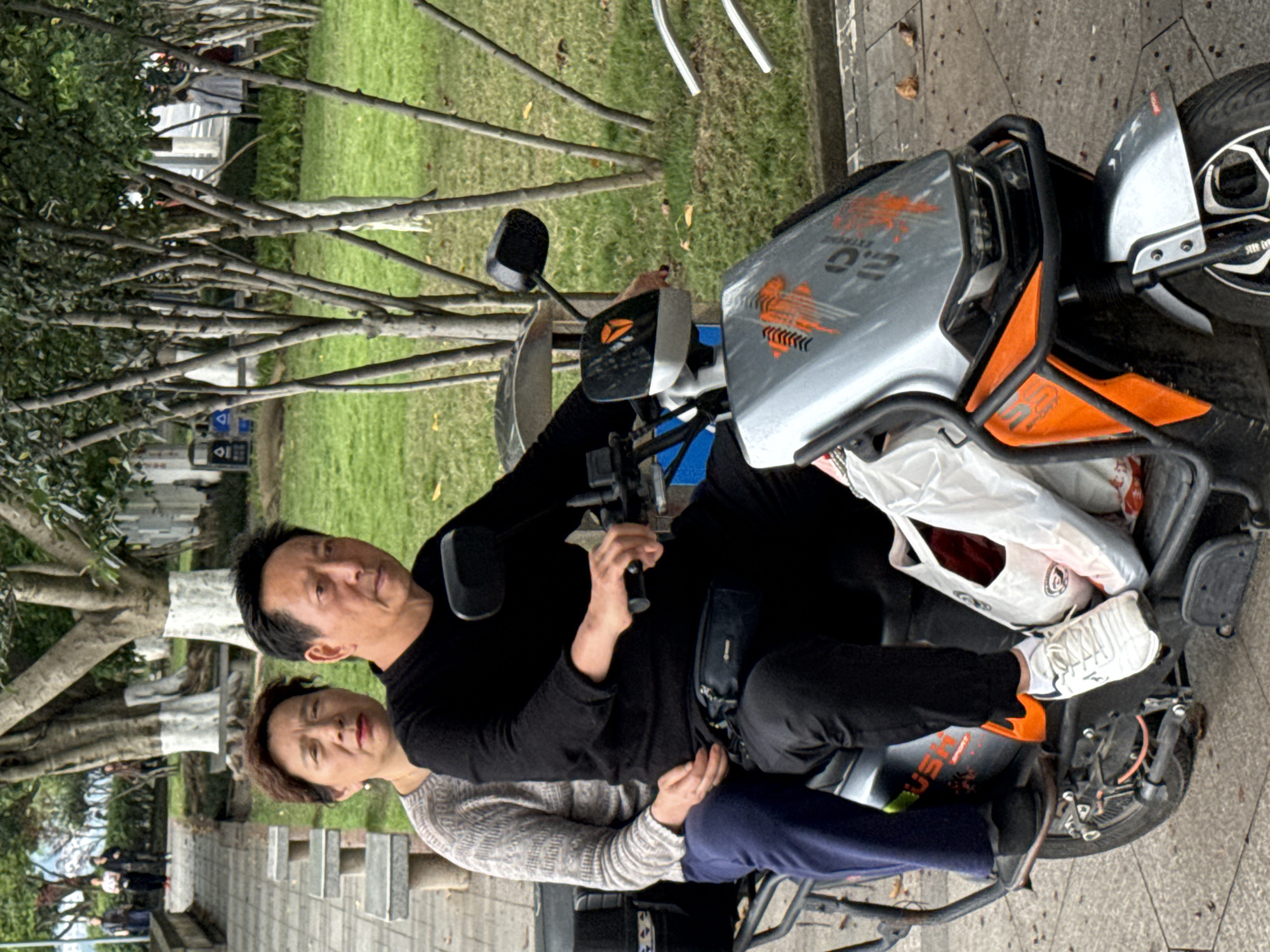 An adult male and adult female sit on a motorized scooter parked on a concrete sidewalk in Jiangjin, China. The man, wearing a black top and black pants, steers the scooter. The woman, wearing a grey sweater and blue pants, sits behind him. The scooter is silver and orange and has a white plastic bag attached. The sidewalk borders a grassy area with trees. A row of park benches runs parallel to the sidewalk.