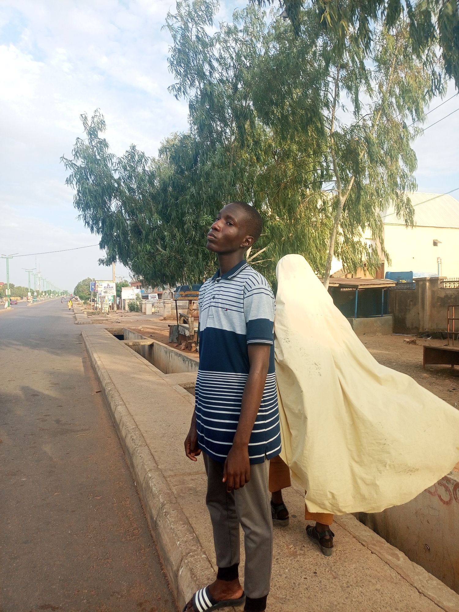 In Tumfure, Nigeria, a young man wearing a horizontally striped navy and grey collared shirt, grey pants, and striped sandals stands at the edge of a sidewalk next to a road. He is looking upwards and to the left. Behind him, another person is holding up a large, light-colored cloth that is billowing in the wind. The road is mostly empty with a few distant cars and streetlights. Buildings and trees are visible in the background.