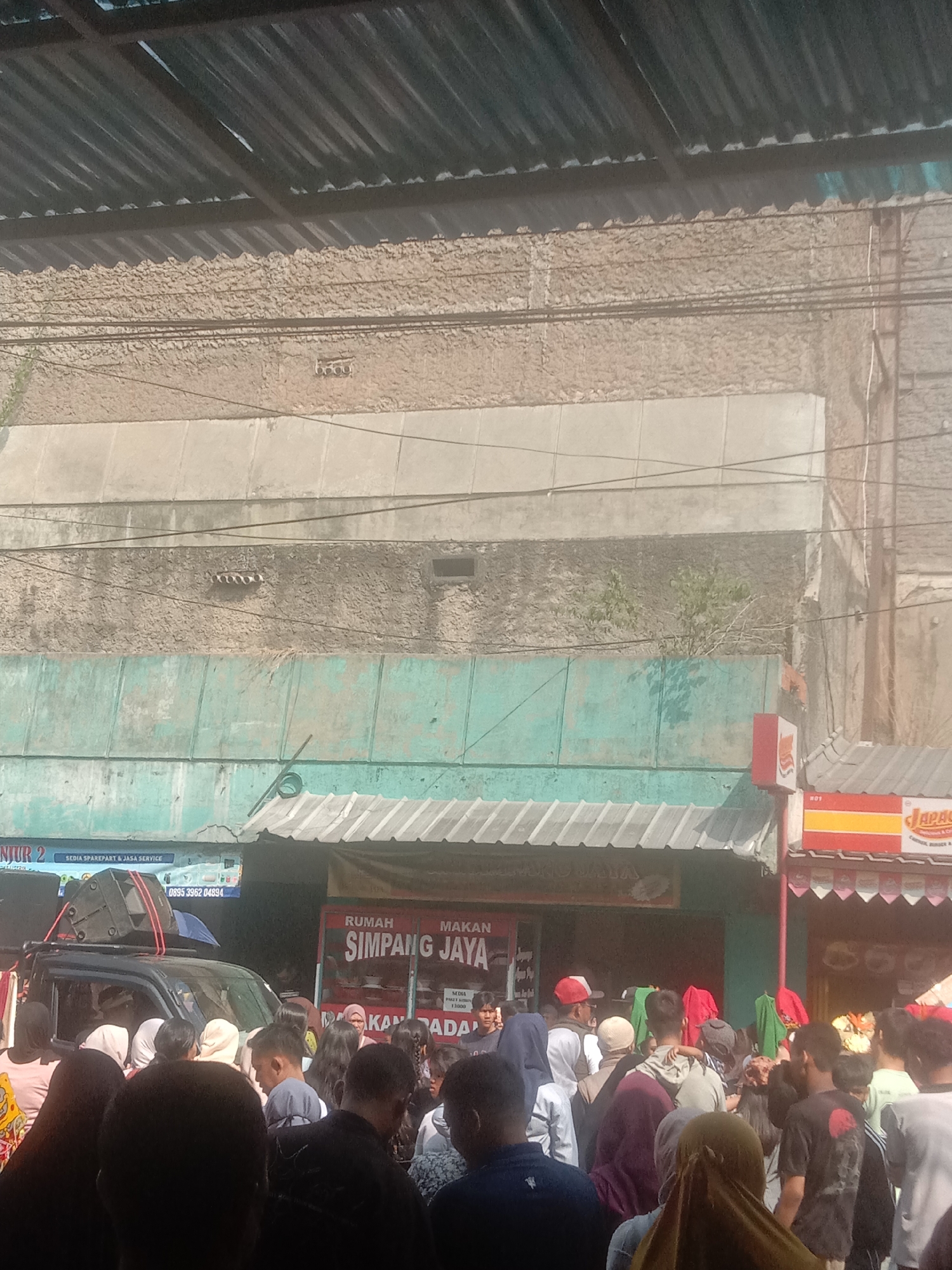 The image depicts a street scene in Cianjur, Indonesia, with a crowd of people gathered in front of several storefronts. A dark-colored vehicle, possibly an SUV, is parked in the foreground, with a large speaker system mounted on its roof. The storefronts appear to be businesses, with signs indicating "RUMAH MAKAN SIMPANG JAYA" and "MAKANAN RADAR." Another sign visible reads "SEDIA SPAREPART & JASA SERVICE" with a phone number. The buildings have a textured, weathered appearance, with some sections painted in turquoise. People in the crowd are dressed in a variety of casual clothing, with some wearing headscarves. The overall atmosphere suggests a lively gathering or market. The background shows a multi-story building with a corrugated metal roof structure extending over the street.