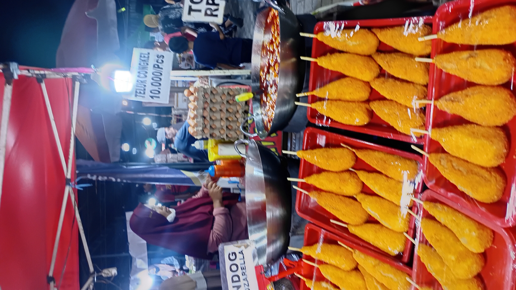This is a daytime, eye-level, medium shot of a food stall at an outdoor market in Jambi, Indonesia. The foreground features several red plastic trays filled with golden-brown fried snacks, possibly corn dogs or similar items coated in breadcrumbs and served on sticks. These snacks are arranged in rows, with some trays covered in plastic wrap.
Behind the snacks, there are two large metal woks or frying pans placed on a dark cooking surface. The wok on the left appears to be empty or contains cooking oil, while the wok on the right is filled with a bubbling, reddish-orange liquid, suggesting food is being cooked or kept warm.
A woman wearing a maroon hijab and a light-colored top is standing behind the stall, reaching for a bottle. She appears to be a vendor. In the background, there are other people, some browsing and others working at adjacent stalls. There are signs with text in Indonesian. One sign clearly visible says "TELUR CONGKEL 10.000/Pcs," indicating a price for a specific food item. Another sign with the letters "NDOG" and "MOZZARELLA" is also visible, suggesting the type of food being sold. The stall is covered by a red canopy, and the lighting suggests it is either late afternoon or the stall is illuminated by artificial lights, creating a slightly blurry effect common in low-light photography. The overall impression is that of a busy, vibrant street food scene in Jambi.