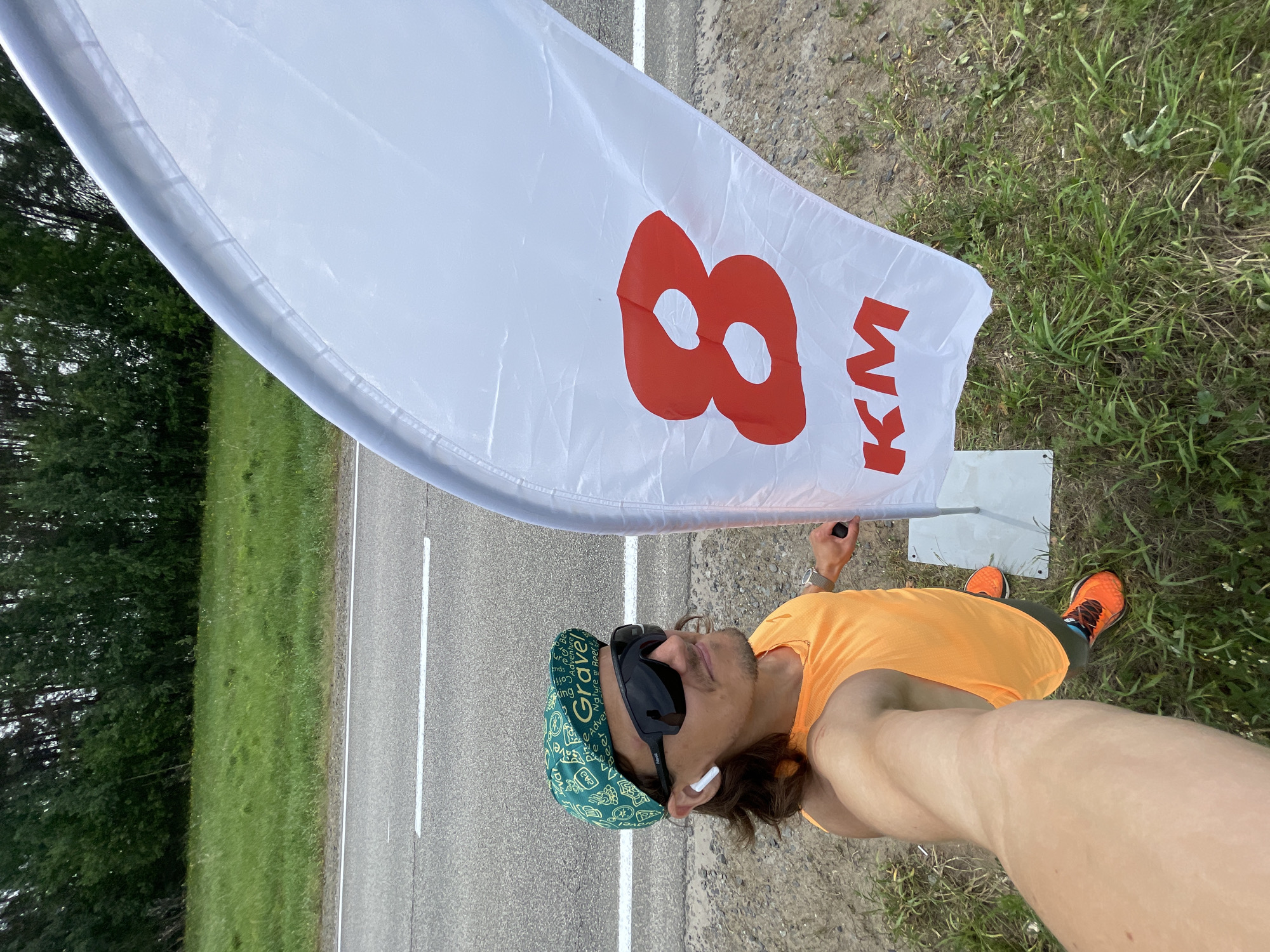 A man is standing on the side of a road, holding a large white flag with "8 KM" printed on it in red. The road is paved and has a white line marking the edge. On the other side of the road is a grassy area and trees. The man is wearing a bright orange shirt, a green patterned hat, and sunglasses. He has an earbud in his ear. He is also wearing orange running shoes. The location is Перевозное, Russia.