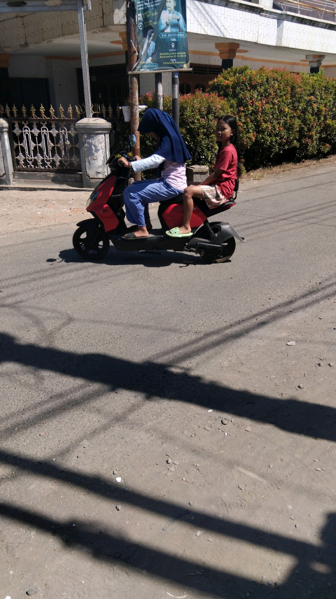 In Sidakaton, Indonesia, two young females are riding on a red scooter along a paved road. The individual in the front, wearing a dark blue hijab and a pink and white shirt with blue pants, is operating the scooter. The passenger, in a red shirt and a patterned skirt, is seated behind. The scene is set against a backdrop of a building with white siding and decorative pillars, and a dense hedge of red-leafed plants. The road surface is asphalt, marked with faint white lines and shadowed by overhead power lines. The overall lighting suggests it is daytime with bright sunlight casting distinct shadows on the road.